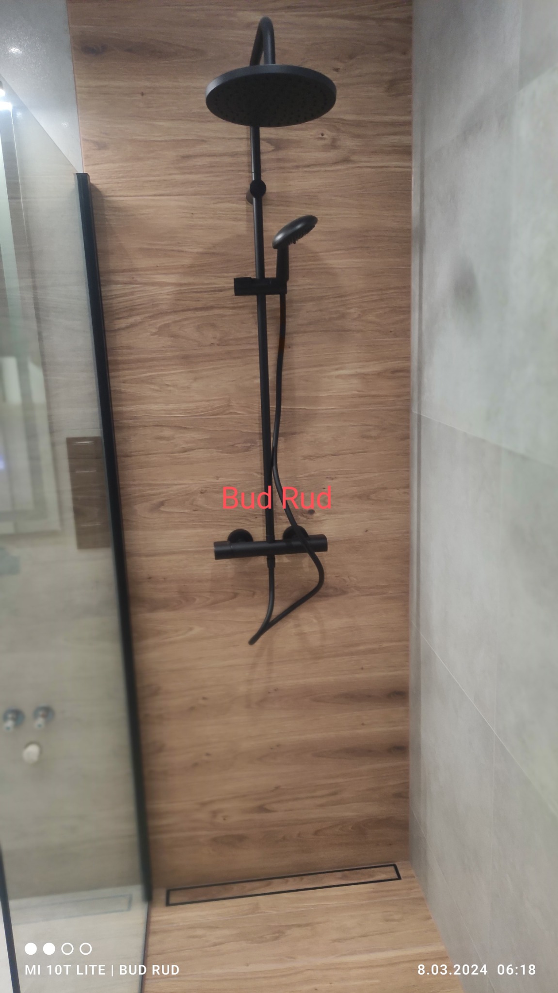 Czarny zestaw prysznicowy z deszczownicą na ścianie wyłożonej panelami imitującymi drewno, widoczny odpływ liniowy w podłodze, kabina prysznicowa z czarnymi profilami po lewej stronie.