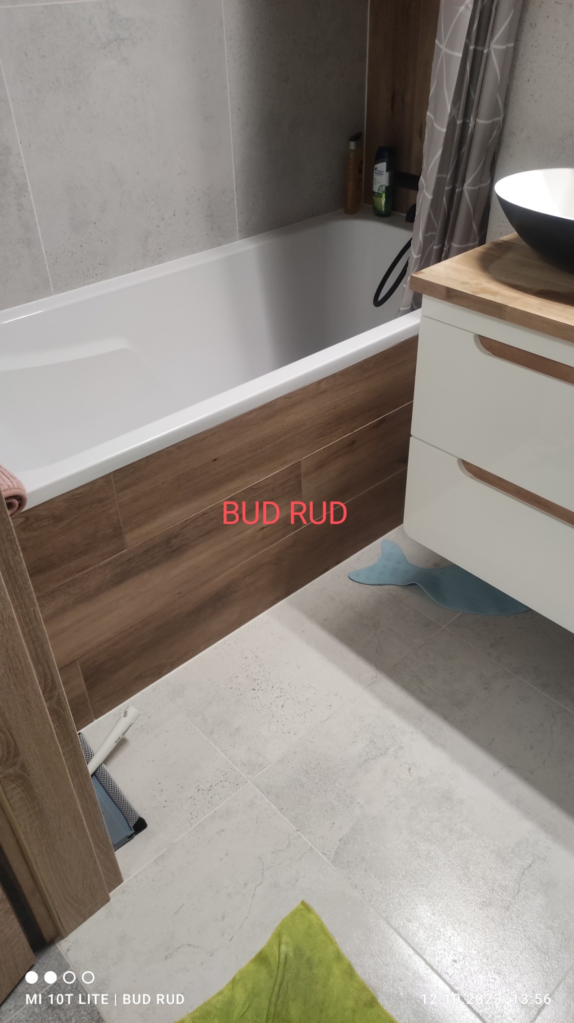 Nowoczesna łazienka z wanną obudowaną panelami imitującymi drewno, szara glazura na ścianach, biała szafka umywalkowa z drewnianym blatem i czarną umywalką, oraz jasnoszare płytki na podłodze.