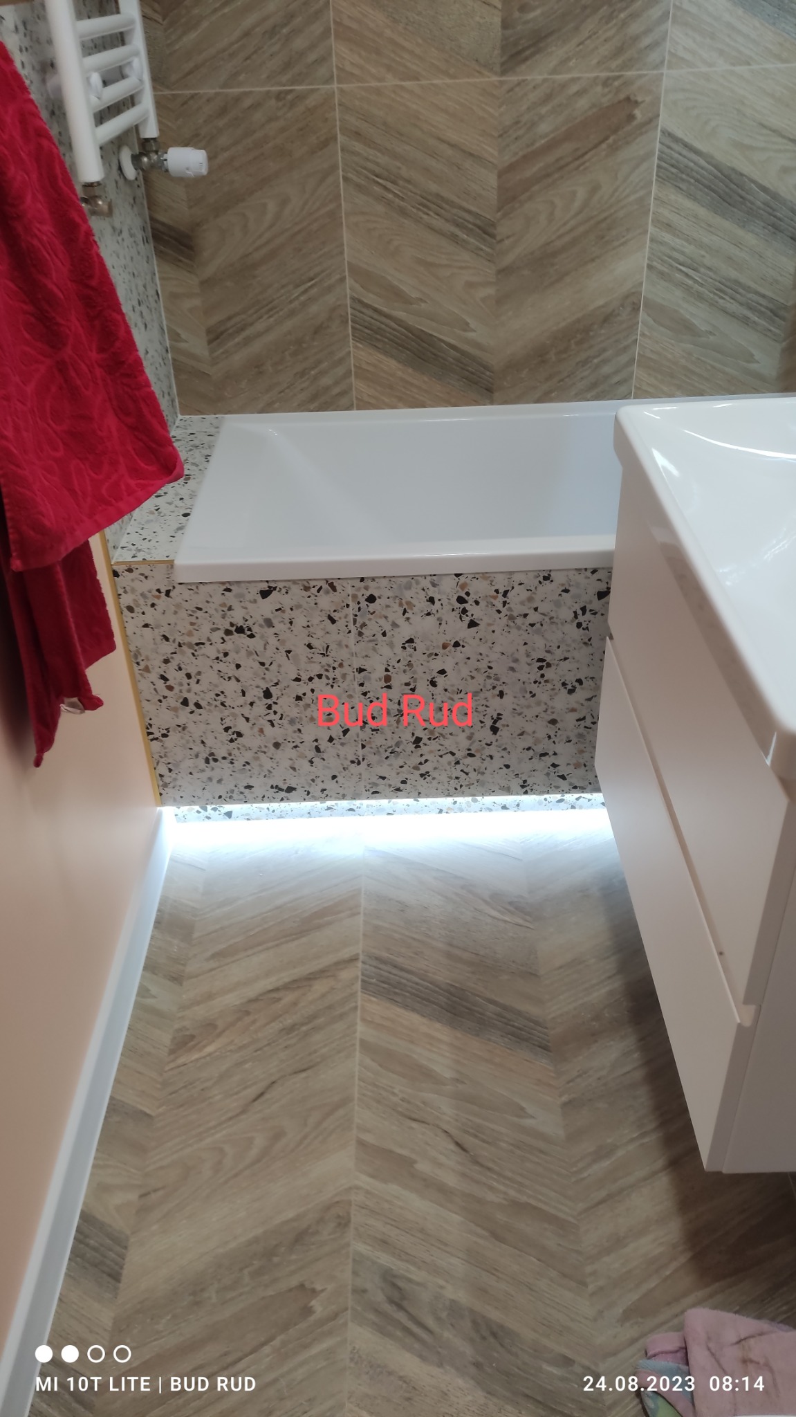 Nowoczesna łazienka z drewnopodobnymi płytkami ułożonymi w jodełkę na podłodze, mozaiką na obudowie wanny i podświetleniem LED u dołu, widoczny fragment czerwonego ręcznika na grzejniku.