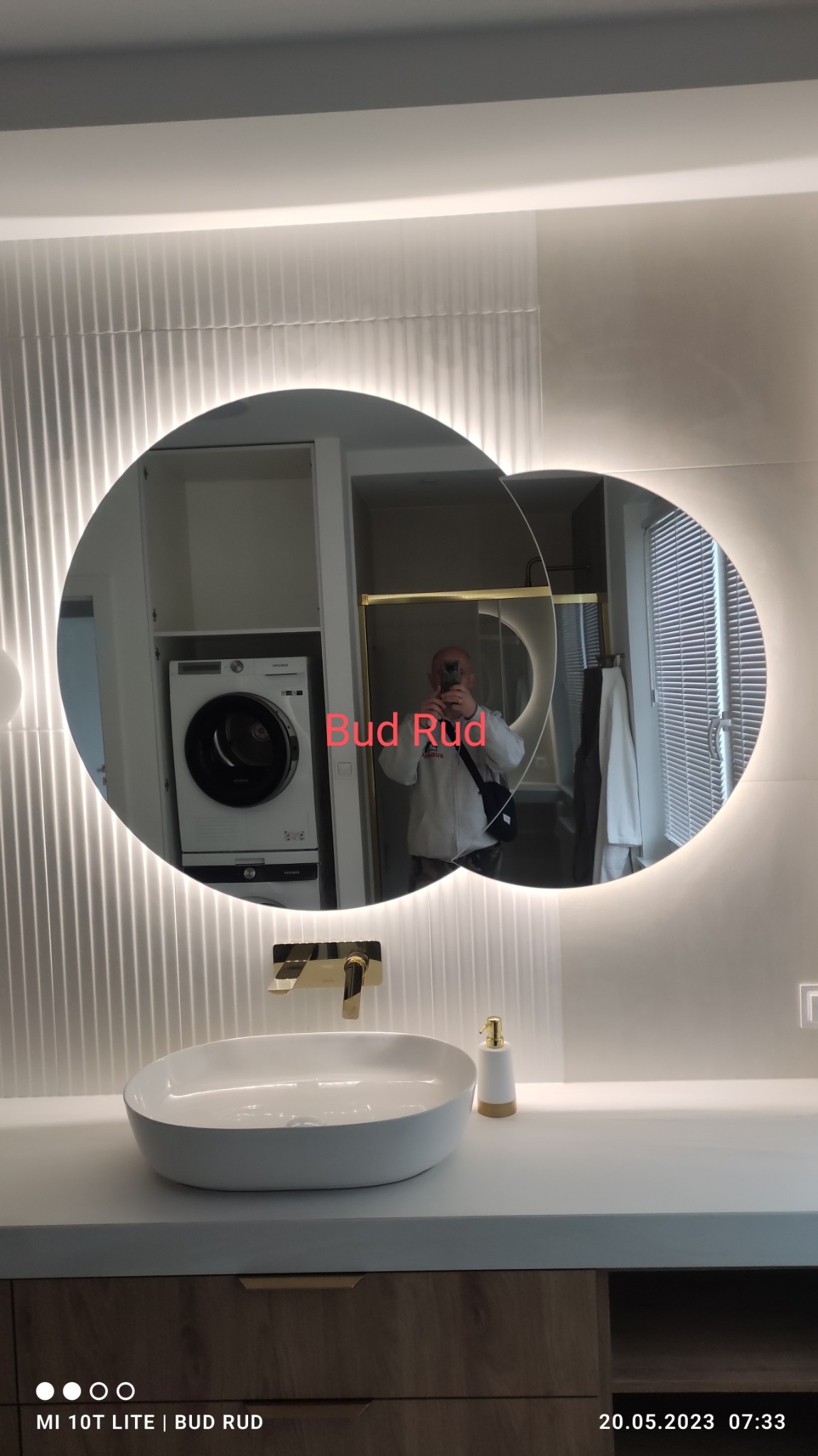 Nowoczesna łazienka z owalną umywalką nablatową, złotą baterią i podświetlanym lustrem; w odbiciu lustra widoczna pralka i suszarka w zabudowie oraz osoba robiąca zdjęcie.