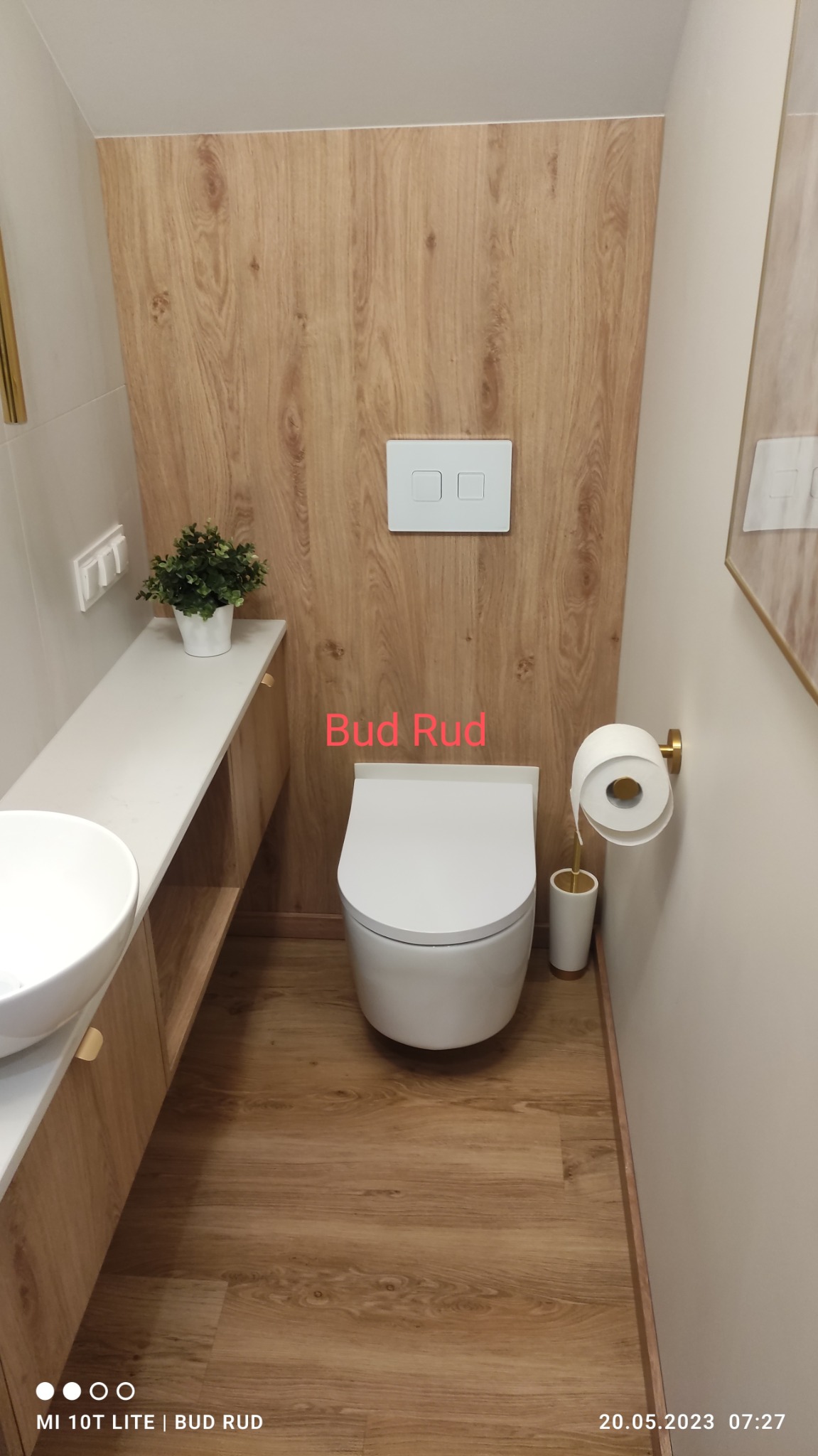 Nowoczesna toaleta z drewnopodobnymi panelami ściennymi i podłogą, biała ceramika sanitarna, wisząca miska WC z przyciskiem spłukującym, złote akcenty w uchwycie na papier toaletowy i doniczce...