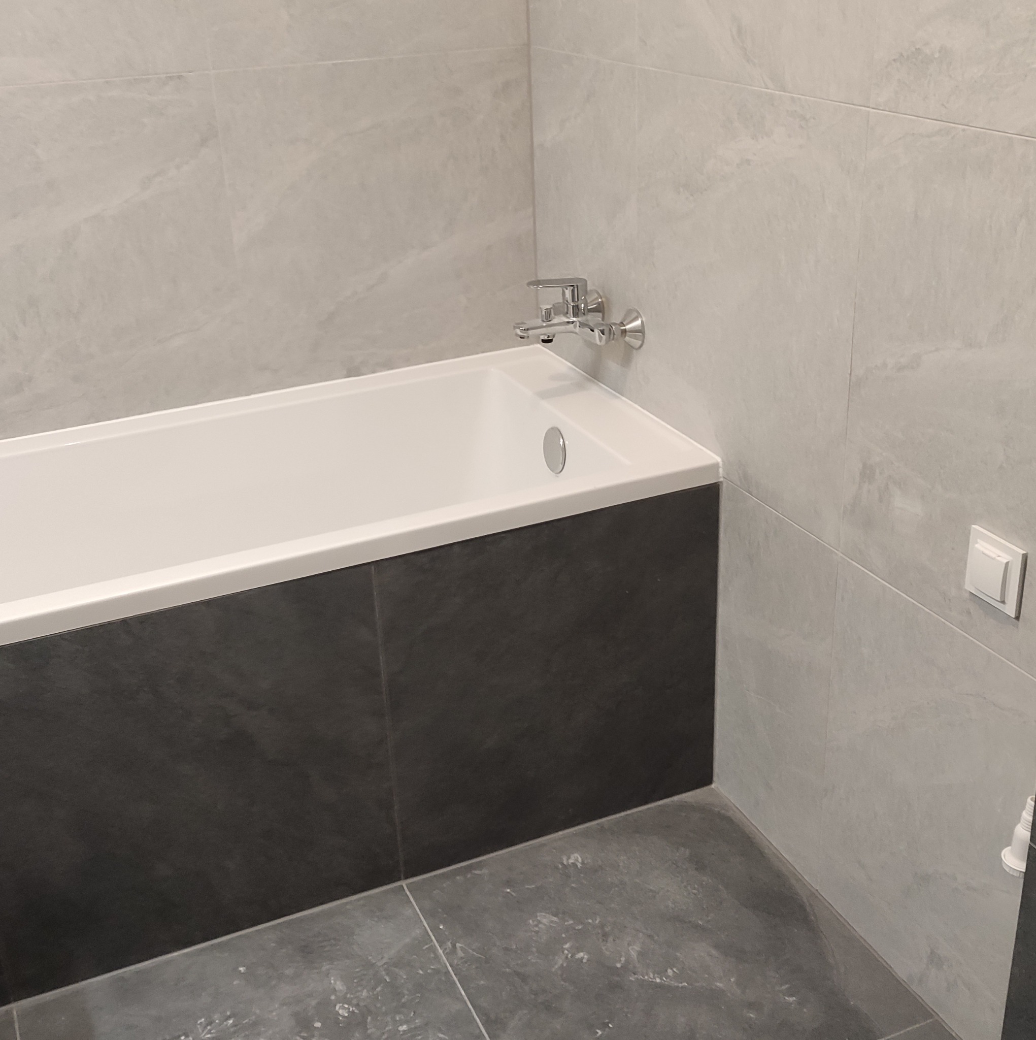 Nowoczesna łazienka z wanną obudowaną ciemnoszarymi płytkami, jasnoszare płytki imitujące marmur na ścianach i podłodze, chromowana bateria wannowa i biały włącznik światła.