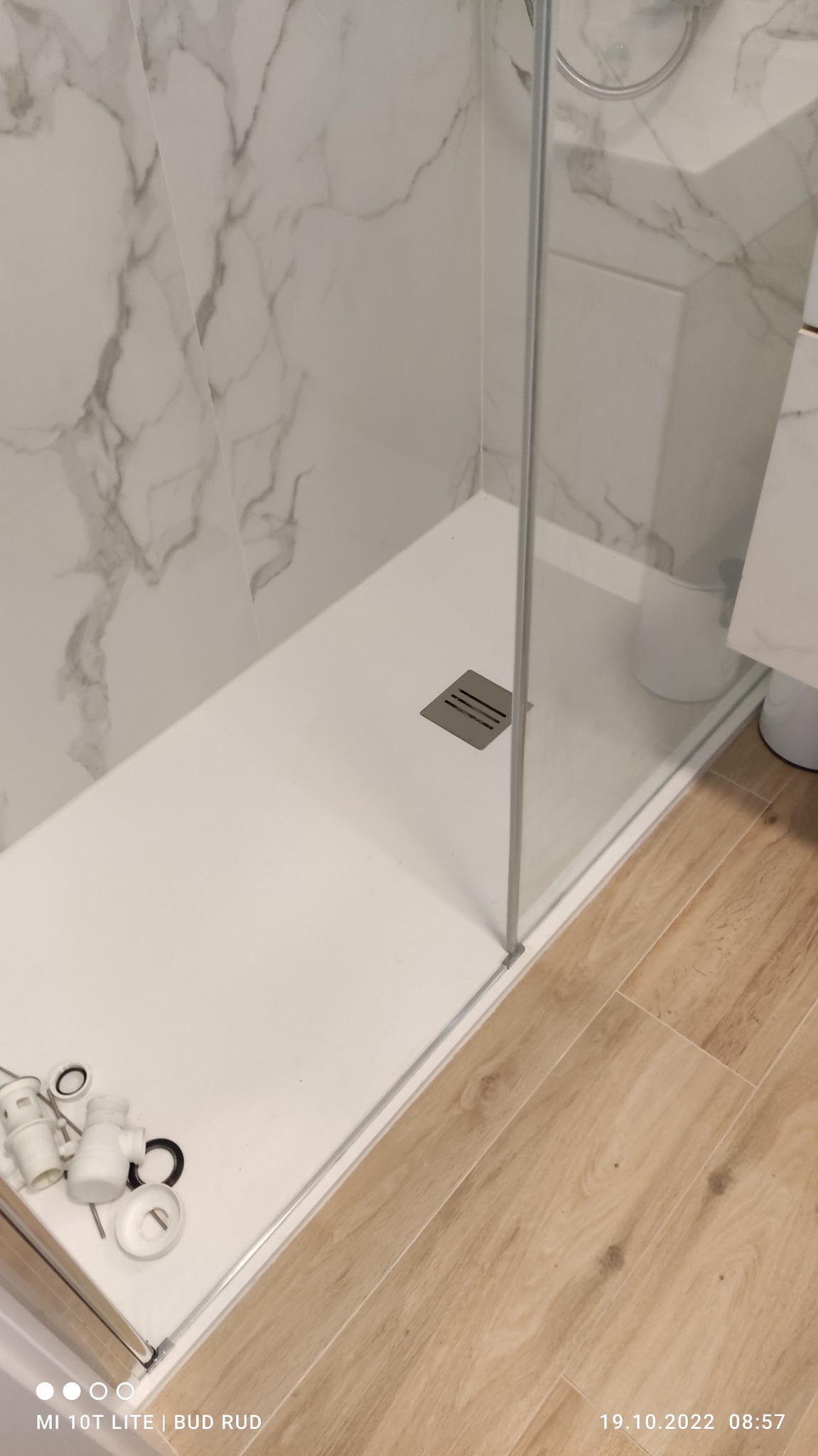 Biały brodzik z odpływem w kabinie prysznicowej z marmurowymi płytkami na ścianie, widoczne elementy montażowe syfonu na brodziku oraz fragment drewnopodobnej podłogi.