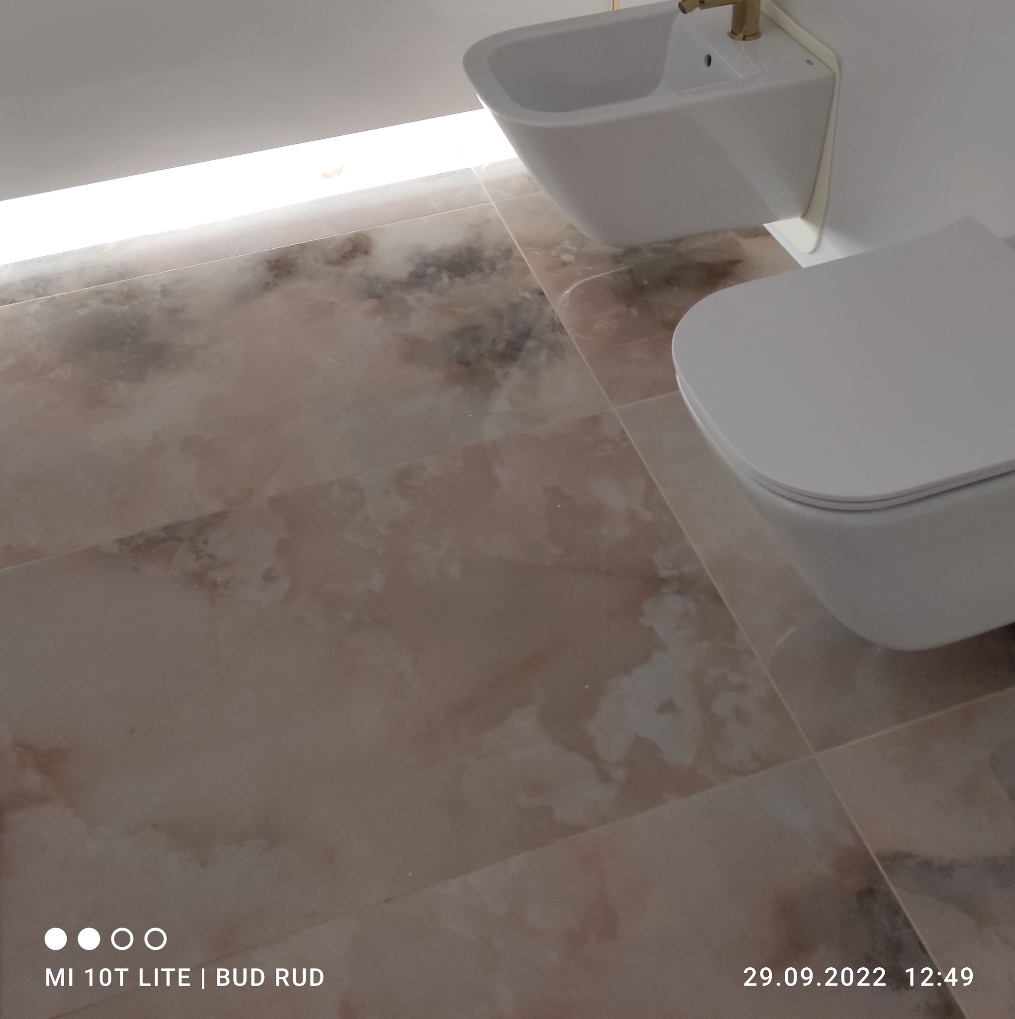 Beżowe płytki podłogowe imitujące marmur w łazience, z widocznym białym bidetem i toaletą podwieszaną, oświetlenie LED pod sufitem.