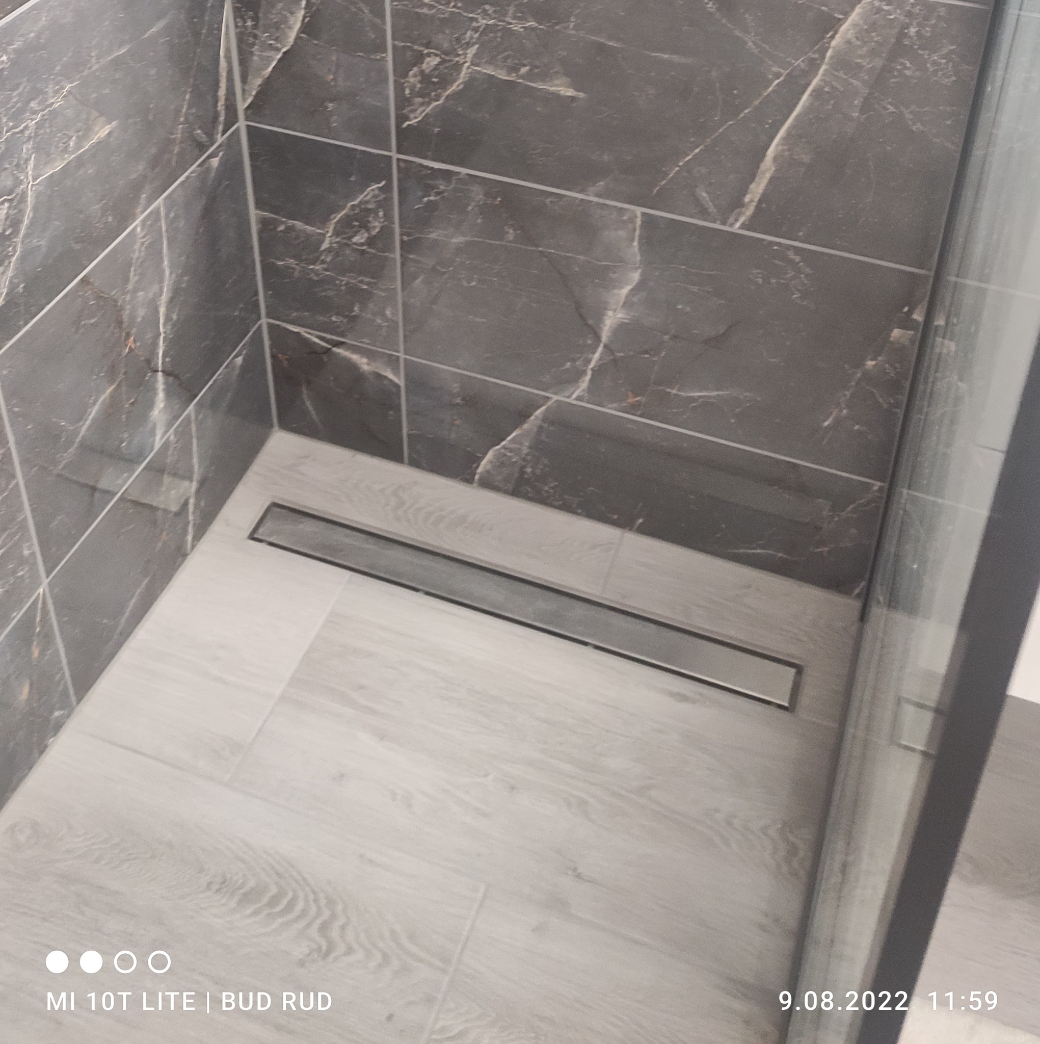 Nowoczesny odpływ liniowy w narożnej kabinie prysznicowej z szarymi płytkami imitującymi marmur na ścianach i jasnymi płytkami podłogowymi imitującymi drewno.