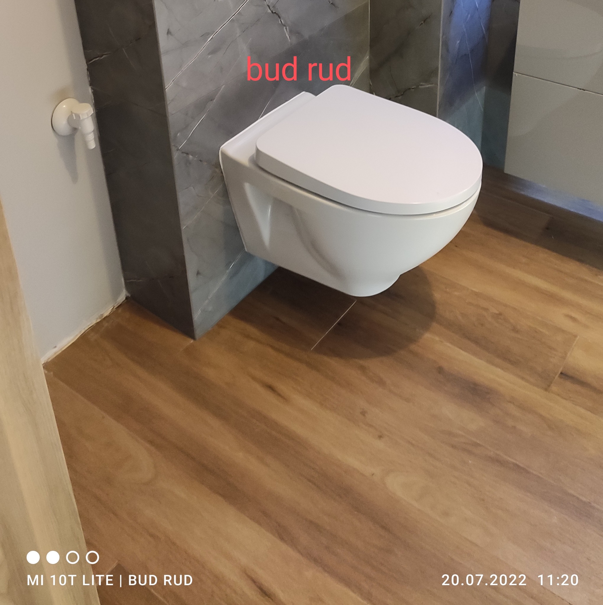 Wisząca toaleta z białą ceramiką, zamontowana na szarej ścianie z marmurowym wzorem, z drewnianą podłogą w tle, widoczny fragment ściany z zaworem.