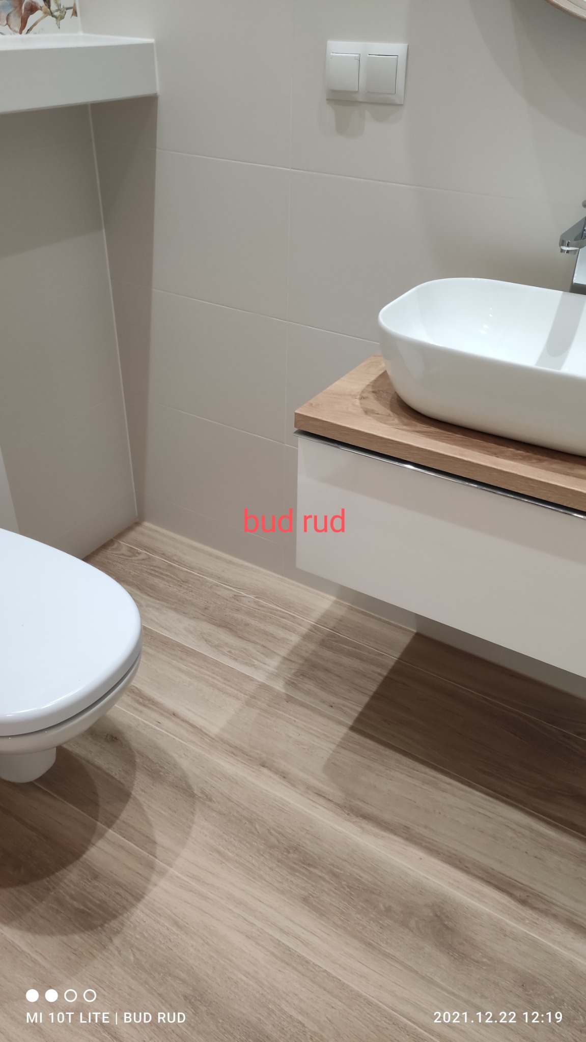 Nowoczesna łazienka z drewnopodobną podłogą, białą umywalką nablatową na szafce z drewnianym blatem i białymi płytkami na ścianach, widoczny fragment toalety.