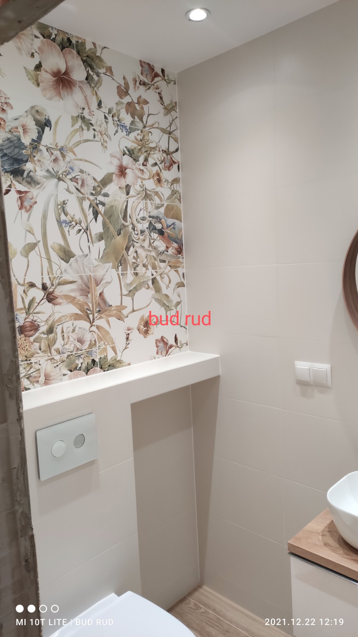 Wykończona łazienka z płytkami w kwiaty i ptaki, podwieszana toaleta, biała umywalka na drewnianym blacie, oświetlenie punktowe.