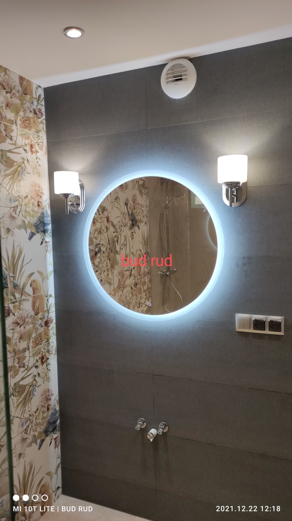 Szara łazienka z okrągłym lustrem podświetlanym LED, wzorzysta tapeta z motywem kwiatowym, płytki imitujące kamień i chromowane baterie.