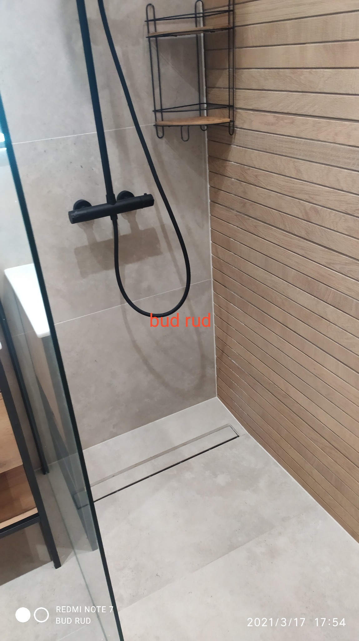 Nowoczesna kabina prysznicowa z czarną armaturą i odpływem liniowym, ściany wyłożone płytkami imitującymi drewno, półka narożna z metalu i drewna.