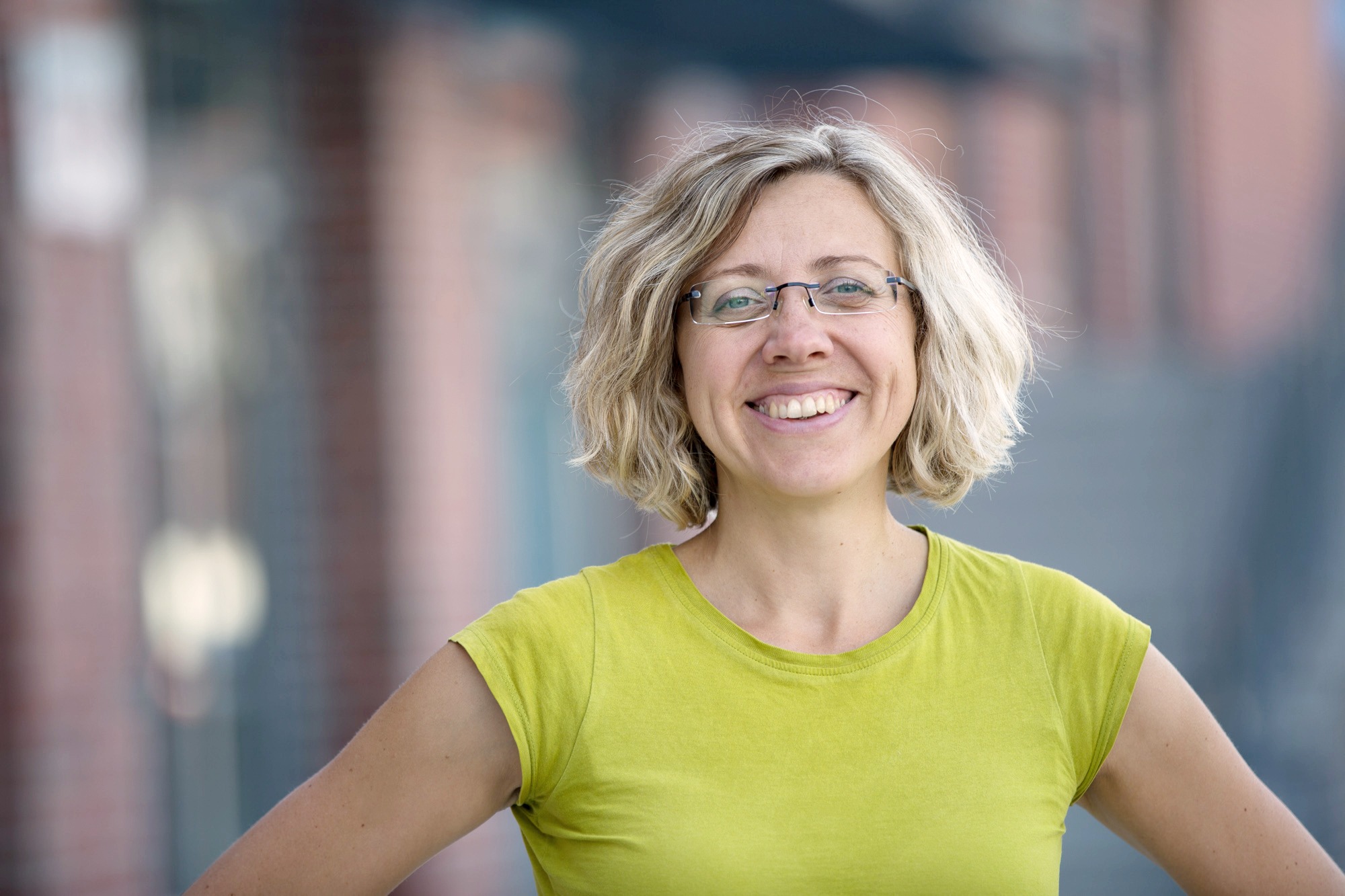 Portret uśmiechniętej kobiety w okularach i zielonej koszulce, z krótkimi blond włosami na rozmytym tle miejskim.