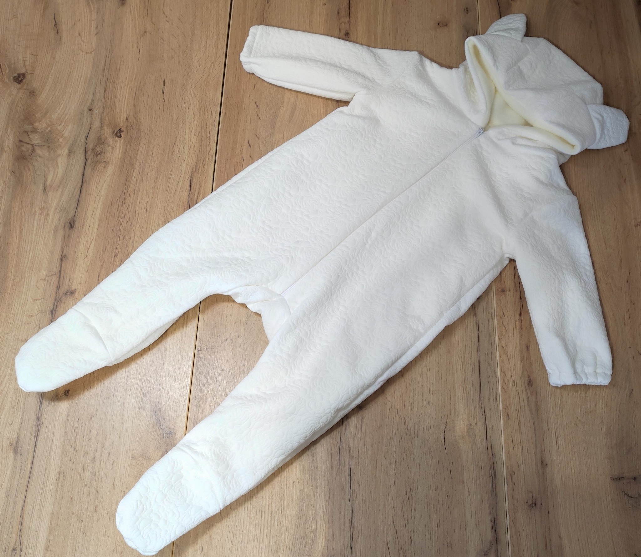 Biały, pluszowy kombinezon dziecięcy z kapturem z uszkami, leżący na drewnianej podłodze.