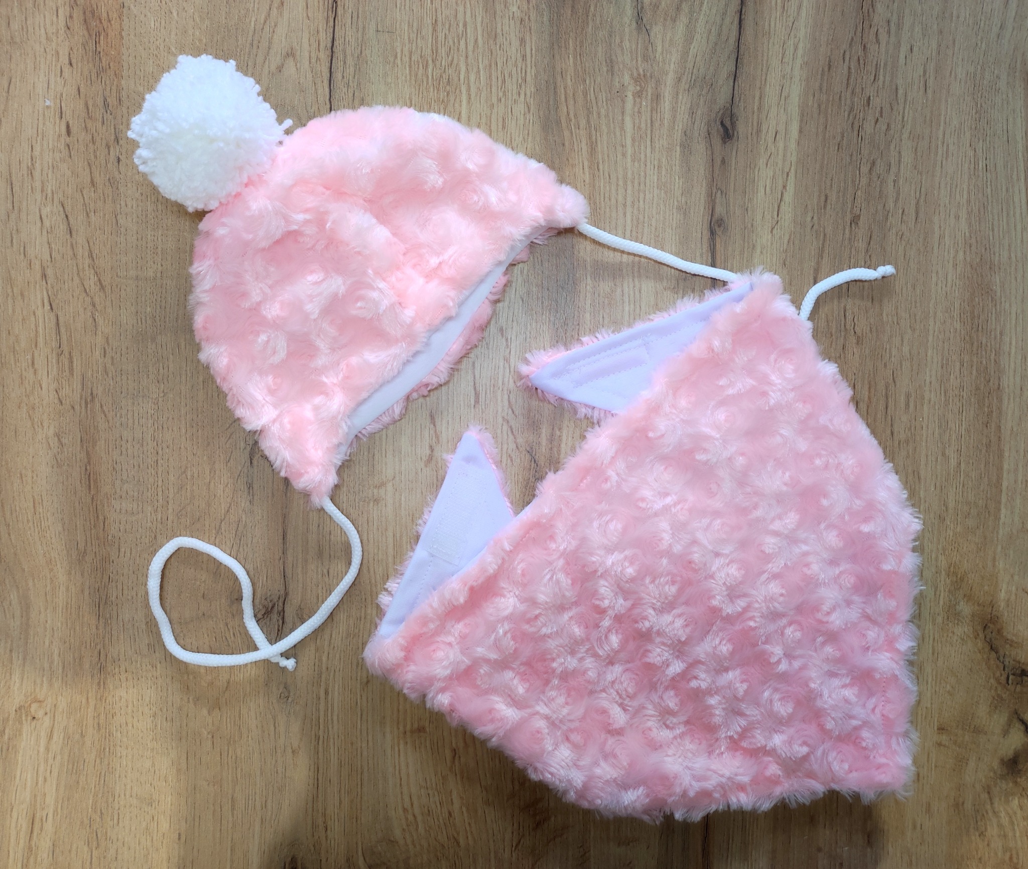 Różowy komplet dziecięcy z miękkiego, wzorzystego materiału: czapka z białym pomponem i wiązaniami oraz trójkątna chusta, leżące na drewnianym podłożu.