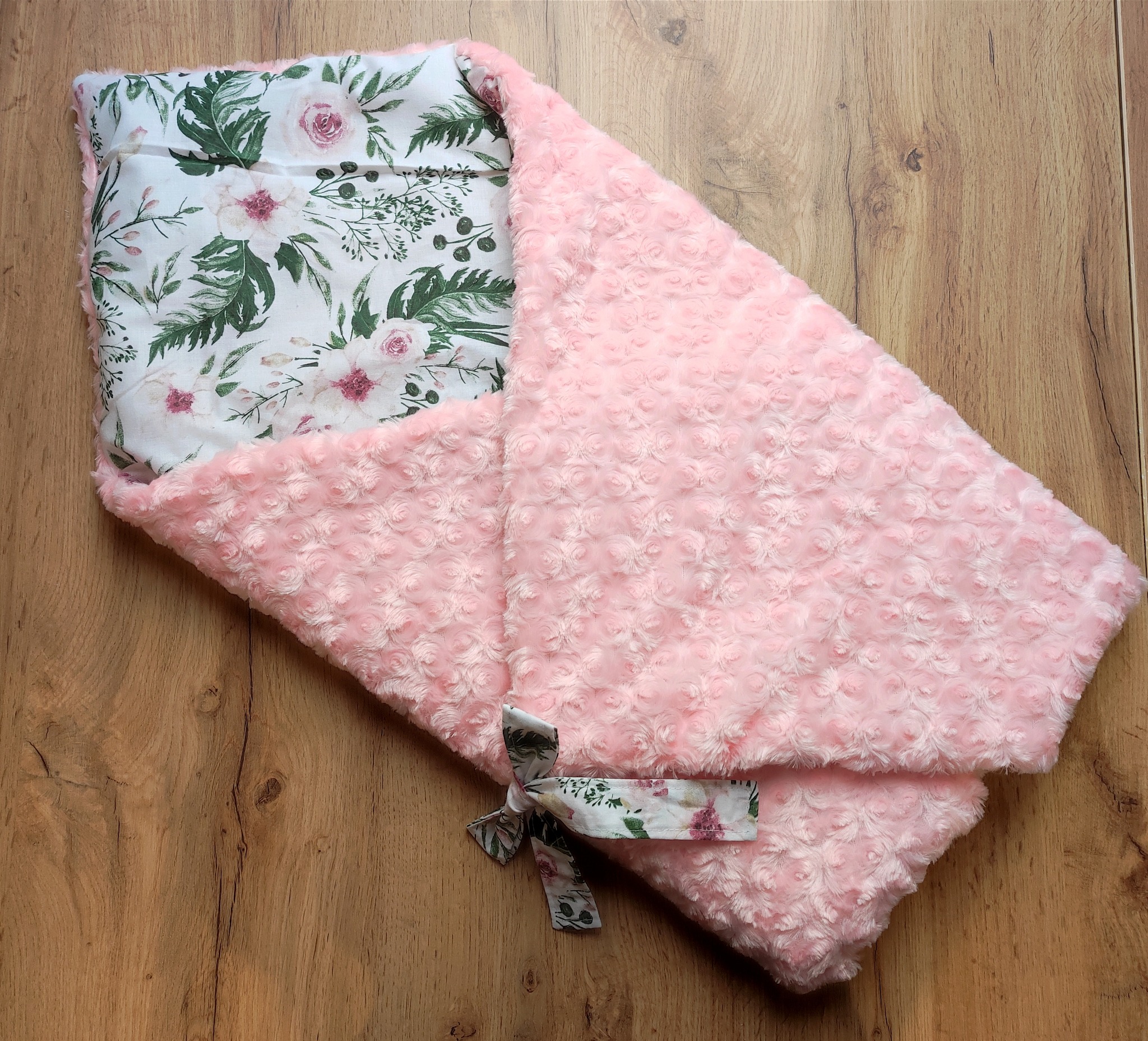 Różowy, pluszowy kocyk niemowlęcy z kwiatowym motywem, zawiązany na kokardkę, leżący na drewnianej podłodze.