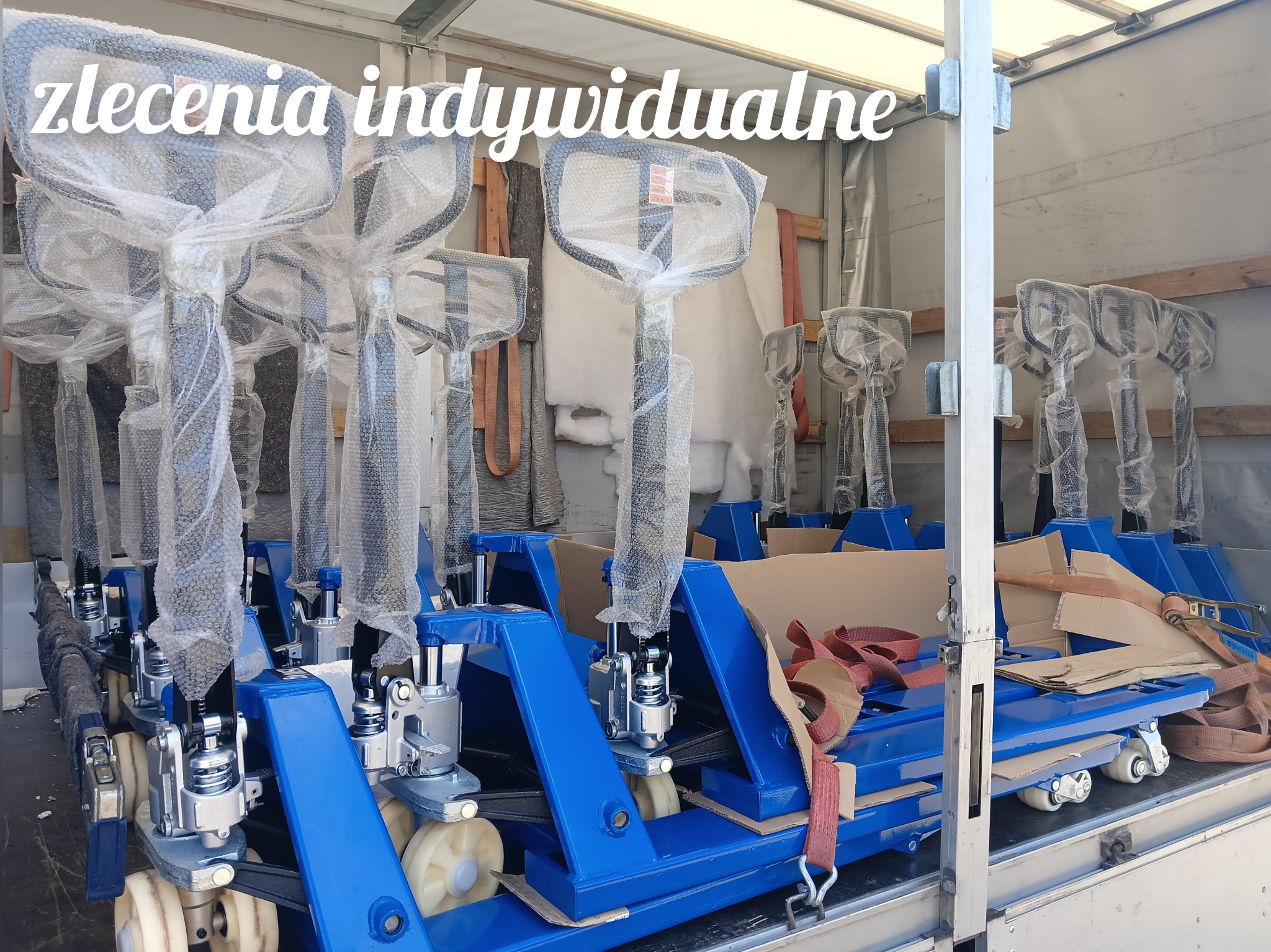 Wnętrze furgonetki wypełnione niebieskimi wózkami transportowymi z platformą, zabezpieczonymi folią bąbelkową i pasami transportowymi.