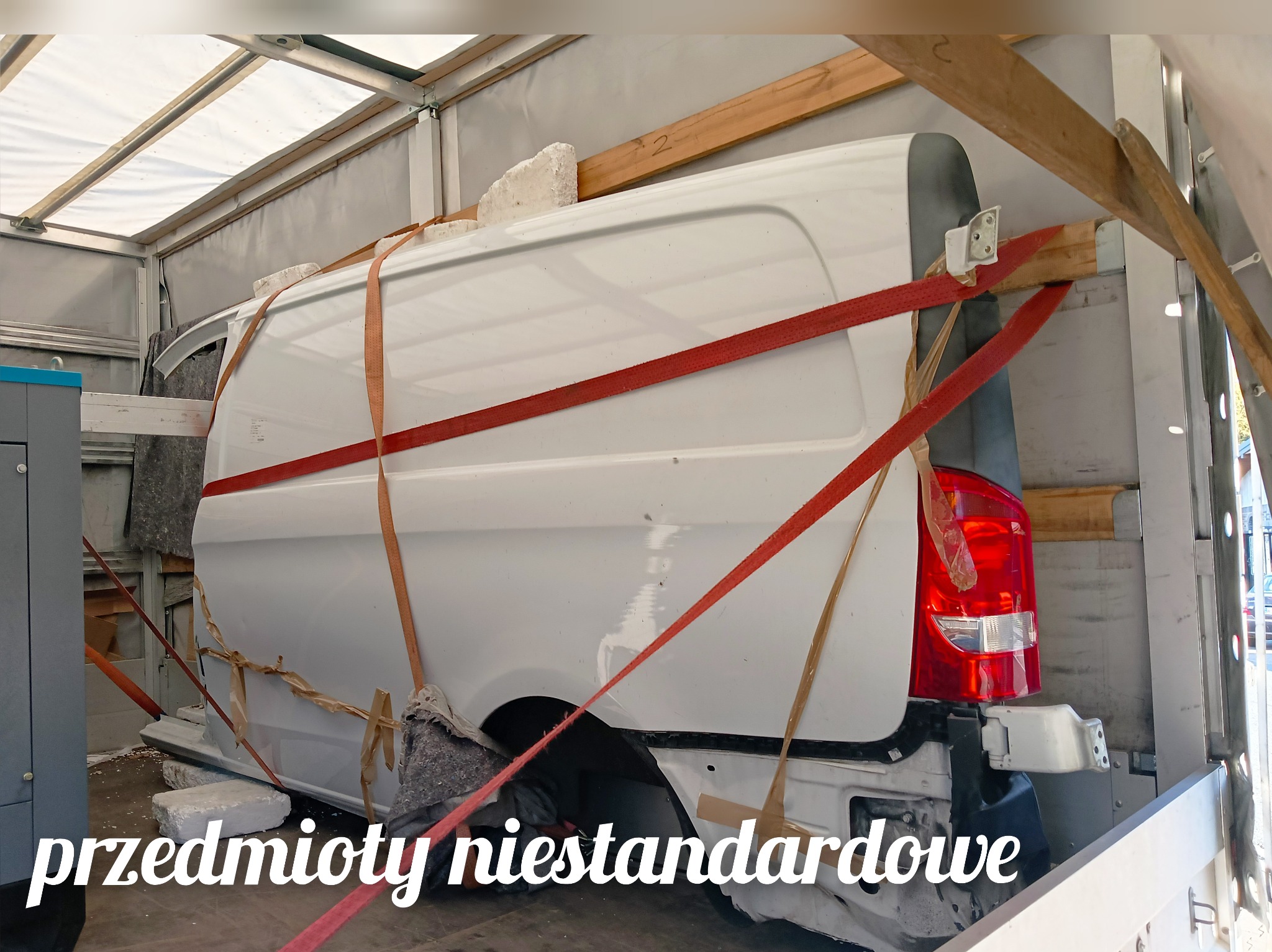 Fragment białego samochodu dostawczego zabezpieczony pasami transportowymi i blokami styropianu wewnątrz przestrzeni ładunkowej, widoczne elementy karoserii i tylna lampa.