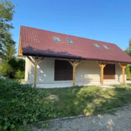 Niewielki, parterowy budynek z szarą elewacją, czerwoną dachówką z trzema oknami dachowymi i drewnianą konstrukcją wspierającą okap, otoczony zielenią.