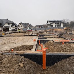 KB PROJEKT KACPER NOWAK - Fundamenty domu w trakcie budowy: wykopane rowy, zbrojenia, rury kanalizacyjne, czarna folia izolacyjna. W tle domy jednorodzinne i sprzęt budowlany.