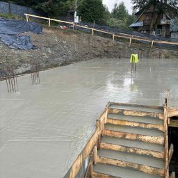 KB PROJEKT KACPER NOWAK - Świeżo wylany, mokry fundament z widocznym szalunkiem schodów i wystającymi prętami zbrojeniowymi na tle ziemi i zabudowań.