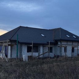 KB PROJEKT KACPER NOWAK - Niedokończony dom z szarej cegły z ciemnym dachem i oknami dachowymi, otoczony suchą trawą, w pochmurny dzień. Widoczne rusztowania i elementy konstrukcyjne.