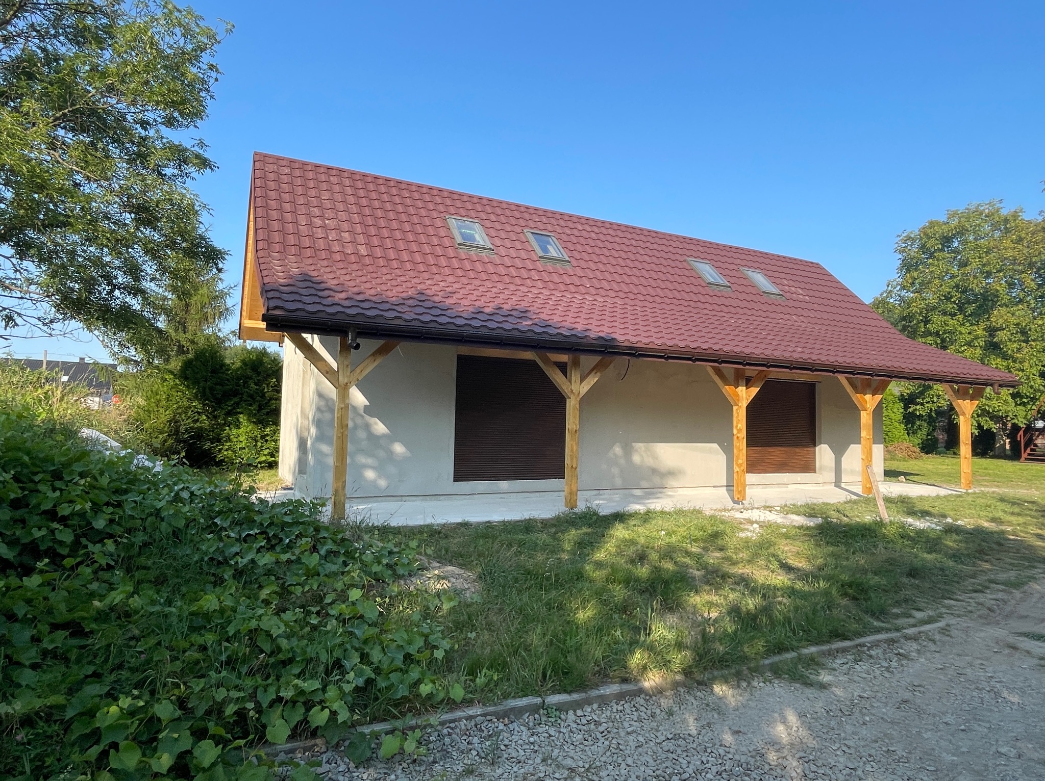 Niewielki, parterowy budynek z szarą elewacją, czerwoną dachówką z trzema oknami dachowymi i drewnianą konstrukcją wspierającą okap, otoczony zielenią.