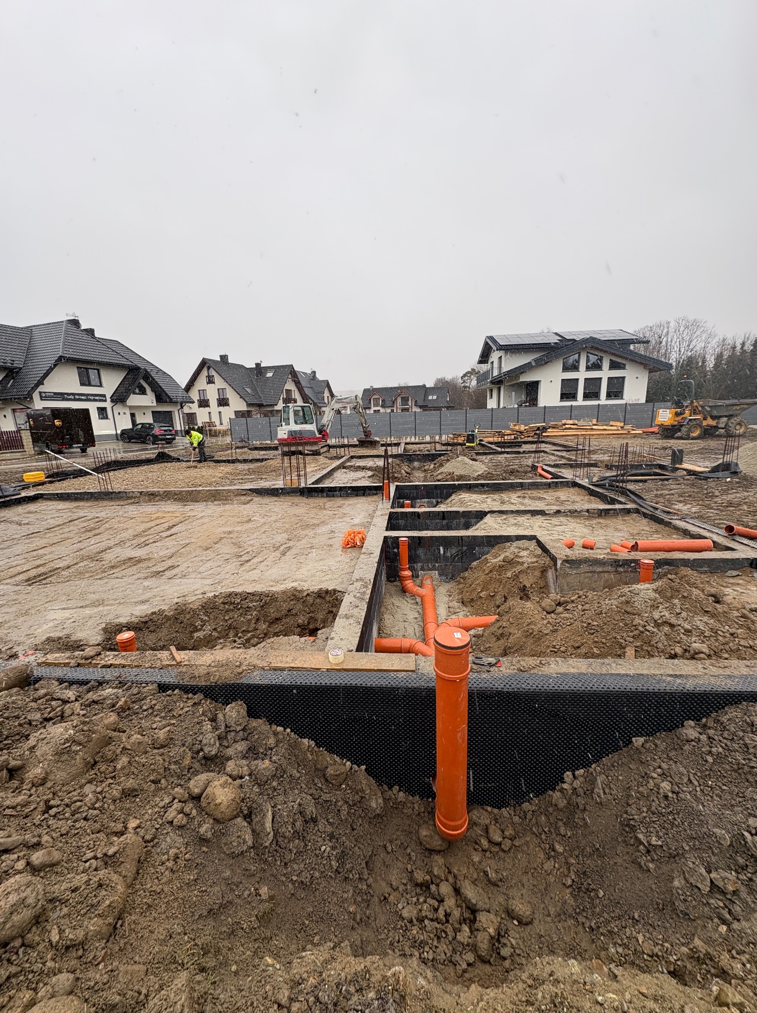 Fundamenty domu w trakcie budowy: wykopane rowy, zbrojenia, rury kanalizacyjne, czarna folia izolacyjna. W tle domy jednorodzinne i sprzęt budowlany.