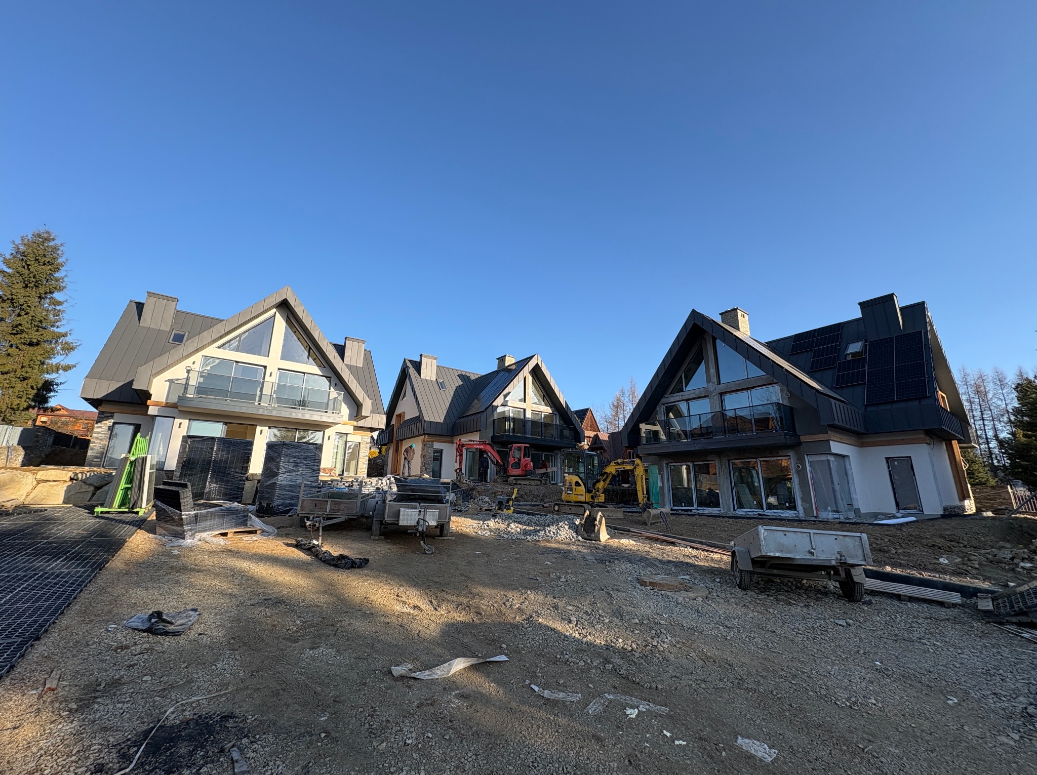 Trzy nowoczesne domy z panelami słonecznymi w trakcie budowy, z widocznymi maszynami budowlanymi i materiałami na placu budowy w słoneczny dzień.