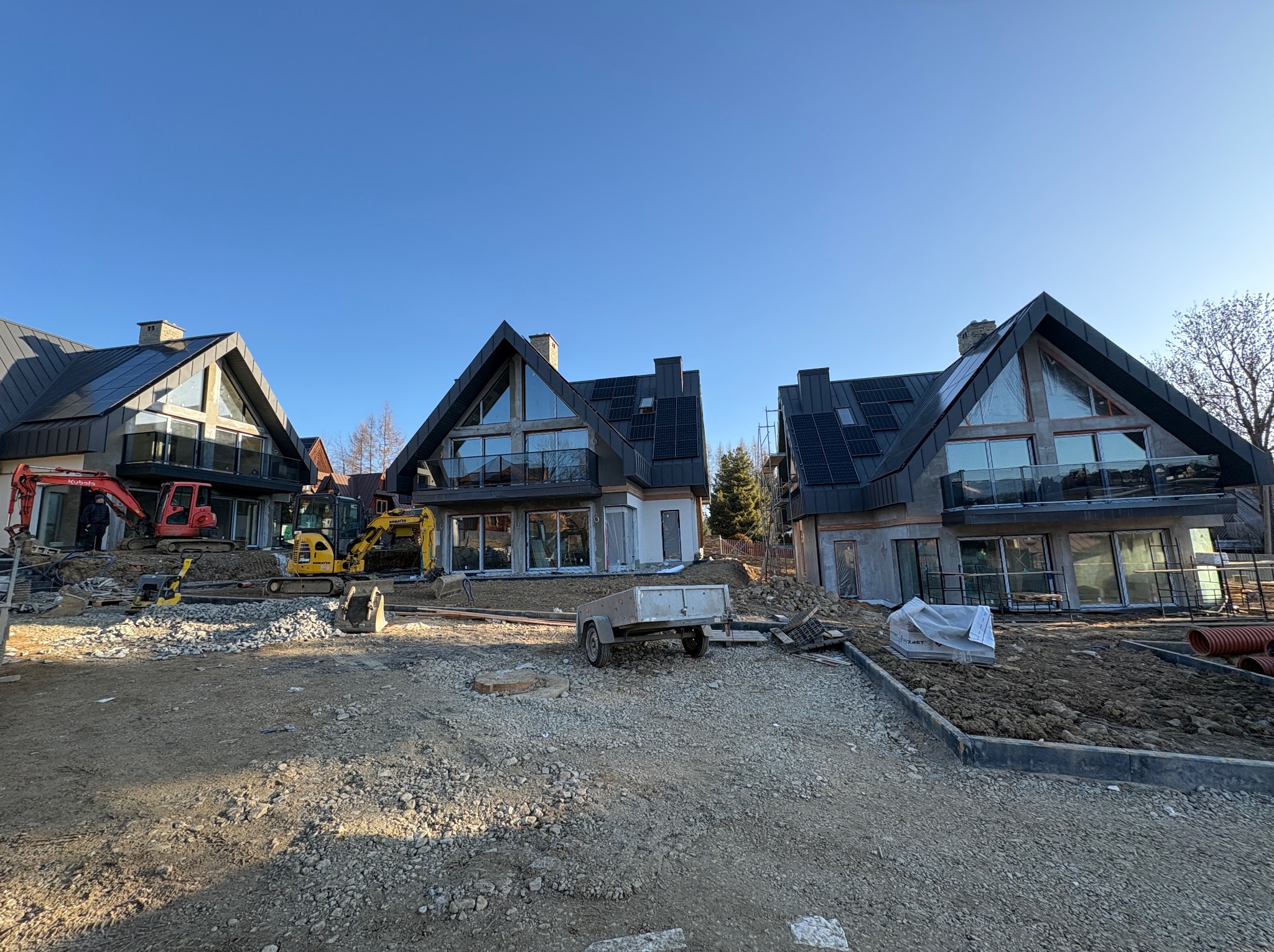 Trzy nowoczesne domy w trakcie budowy, z panelami słonecznymi na dachu i dużymi oknami, na placu budowy z maszynami i materiałami budowlanymi w słoneczny dzień.