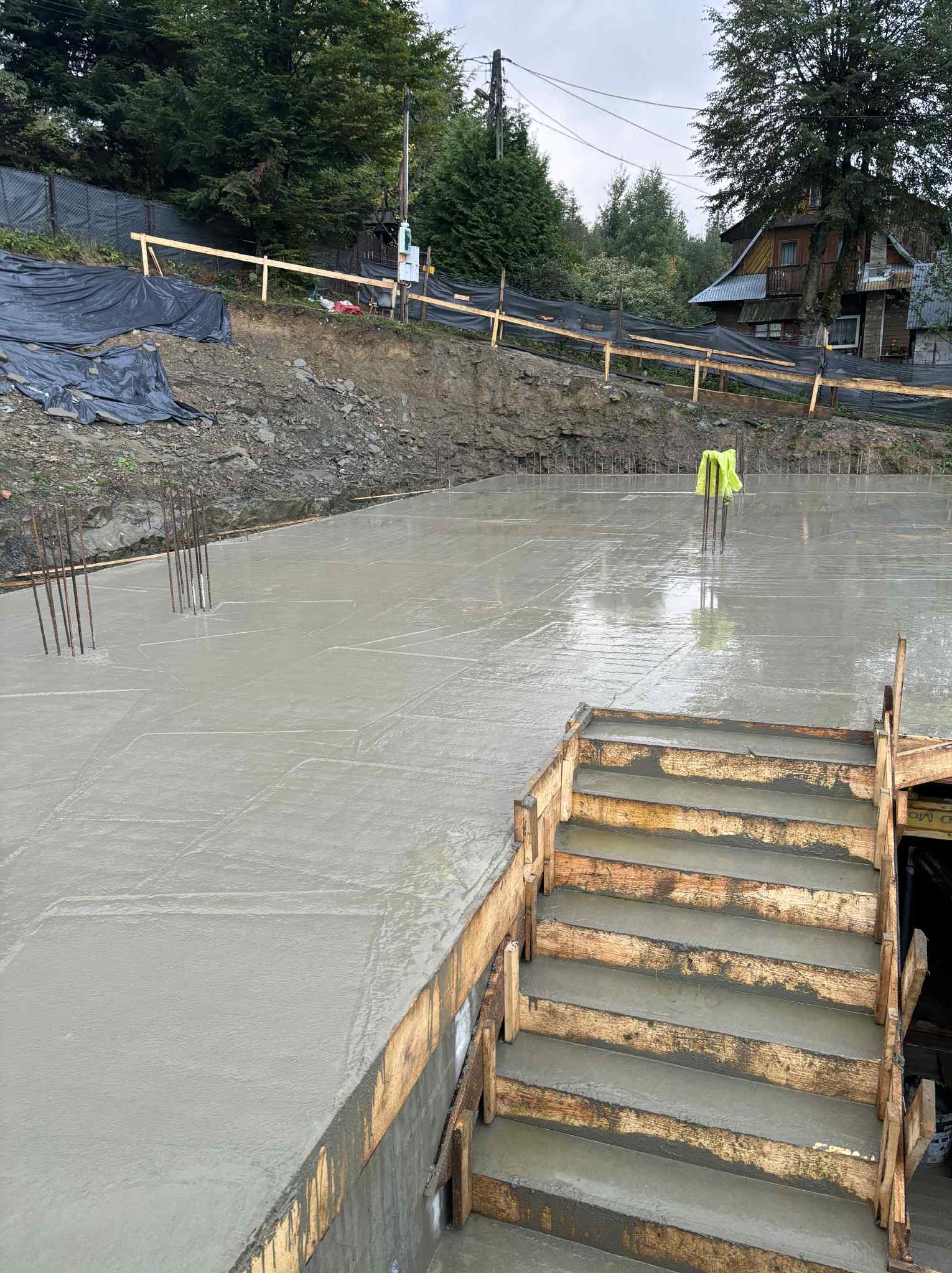 Świeżo wylany, mokry fundament z widocznym szalunkiem schodów i wystającymi prętami zbrojeniowymi na tle ziemi i zabudowań.