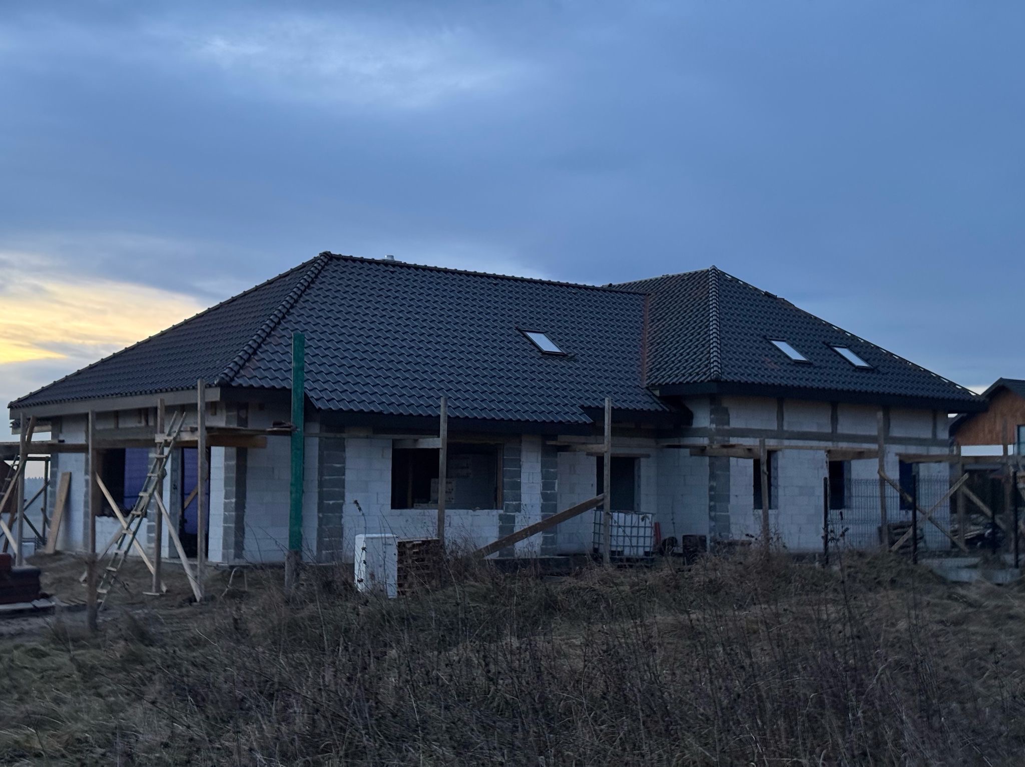 Niedokończony dom z szarej cegły z ciemnym dachem i oknami dachowymi, otoczony suchą trawą, w pochmurny dzień. Widoczne rusztowania i elementy konstrukcyjne.