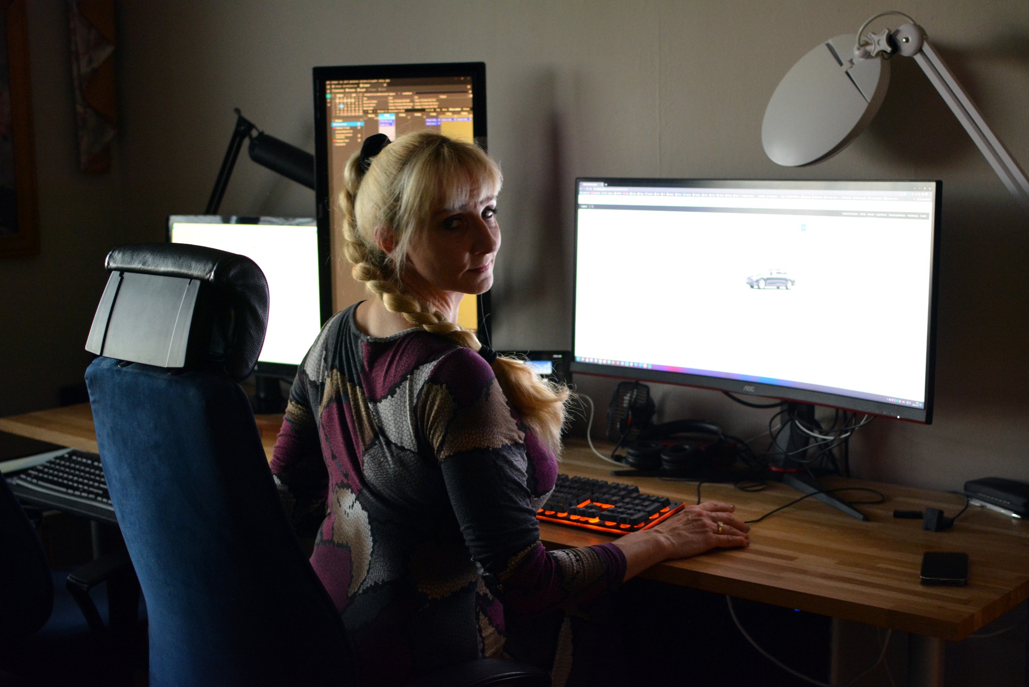 Kobieta z blond warkoczem spogląda przez ramię siedząc przed trzema monitorami komputerowymi na drewnianym biurku, oświetlonym lampą z okrągłym kloszem.