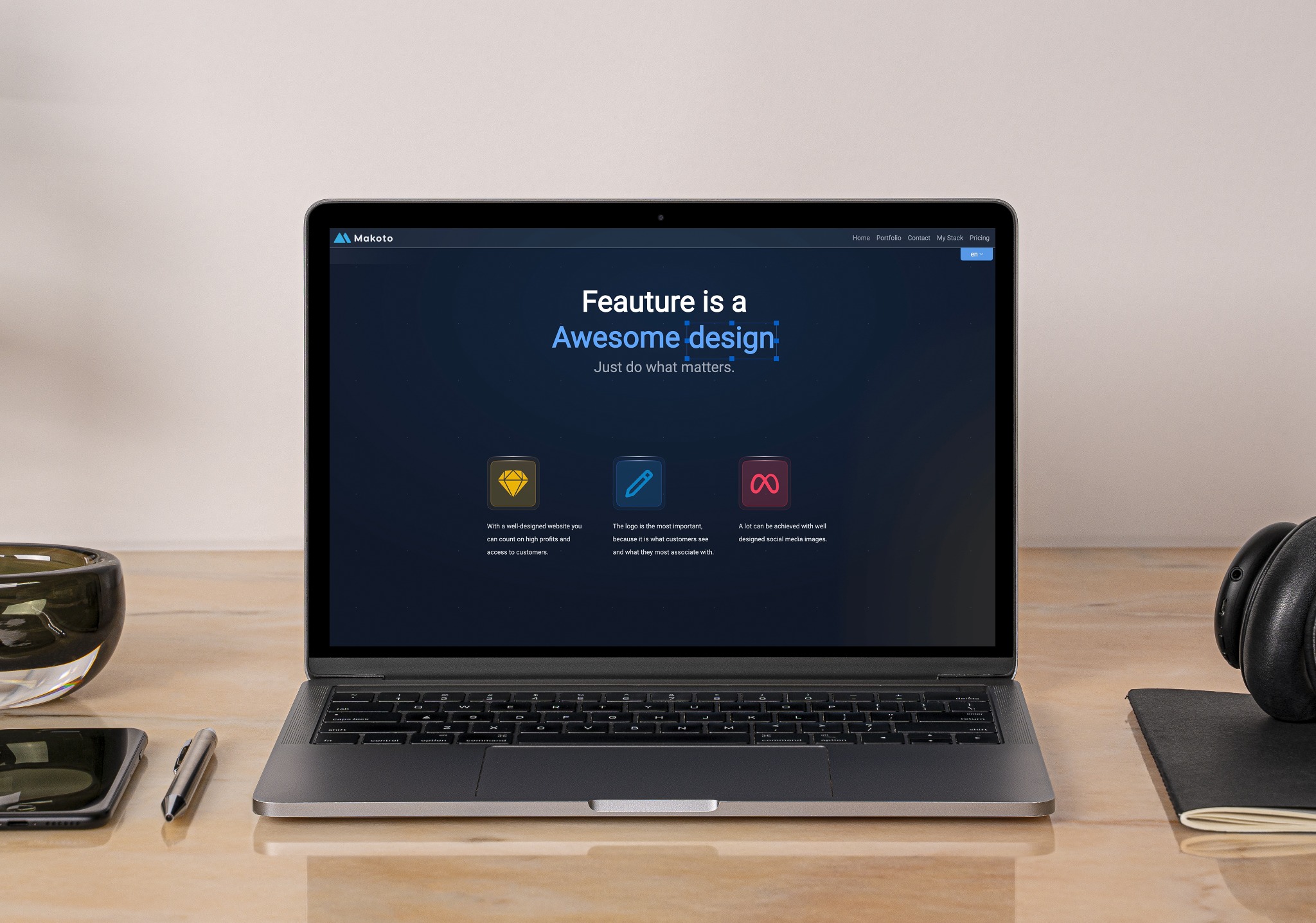 Ekran laptopa prezentujący stronę internetową z hasłem 'Feauture is a Awesome design' i ikonami diamentu, ołówka i nieskończoności, obok telefon, długopis, słuchawki i notatnik na jasnym biurku.