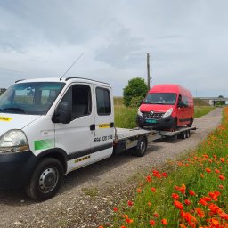 Mieczysław wojtysiak Auto pomoc - Biała pomoc drogowa Renault Master z czerwoną furgonetką na platformie, stojąca na żwirowej drodze wśród kwitnących maków, z mostem i torami kolejowymi w tle.