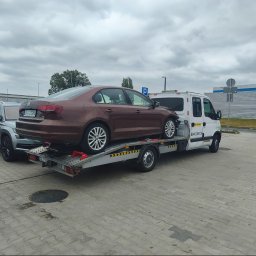 Mieczysław wojtysiak Auto pomoc - Brązowy Volkswagen Jetta na platformie białej lawety z numerem telefonu 694 335 136, zaparkowanej na brukowanym placu pod pochmurnym niebem.