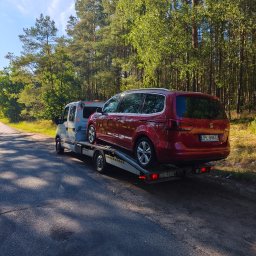 Mieczysław wojtysiak Auto pomoc - Biały pojazd pomocy drogowej transportujący czerwony samochód osobowy SEAT Alhambra na platformie, na tle słonecznego lasu przy drodze, widok z boku.