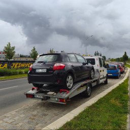 Mieczysław wojtysiak Auto pomoc - Czarny hatchback Toyota Auris z polskimi tablicami ZGL 1596A, załadowany tyłem na platformę transportową, ciągnięty przez biały van. Scena na miejskiej ulicy z graffiti 'DLA JEDNYCH' w tle.