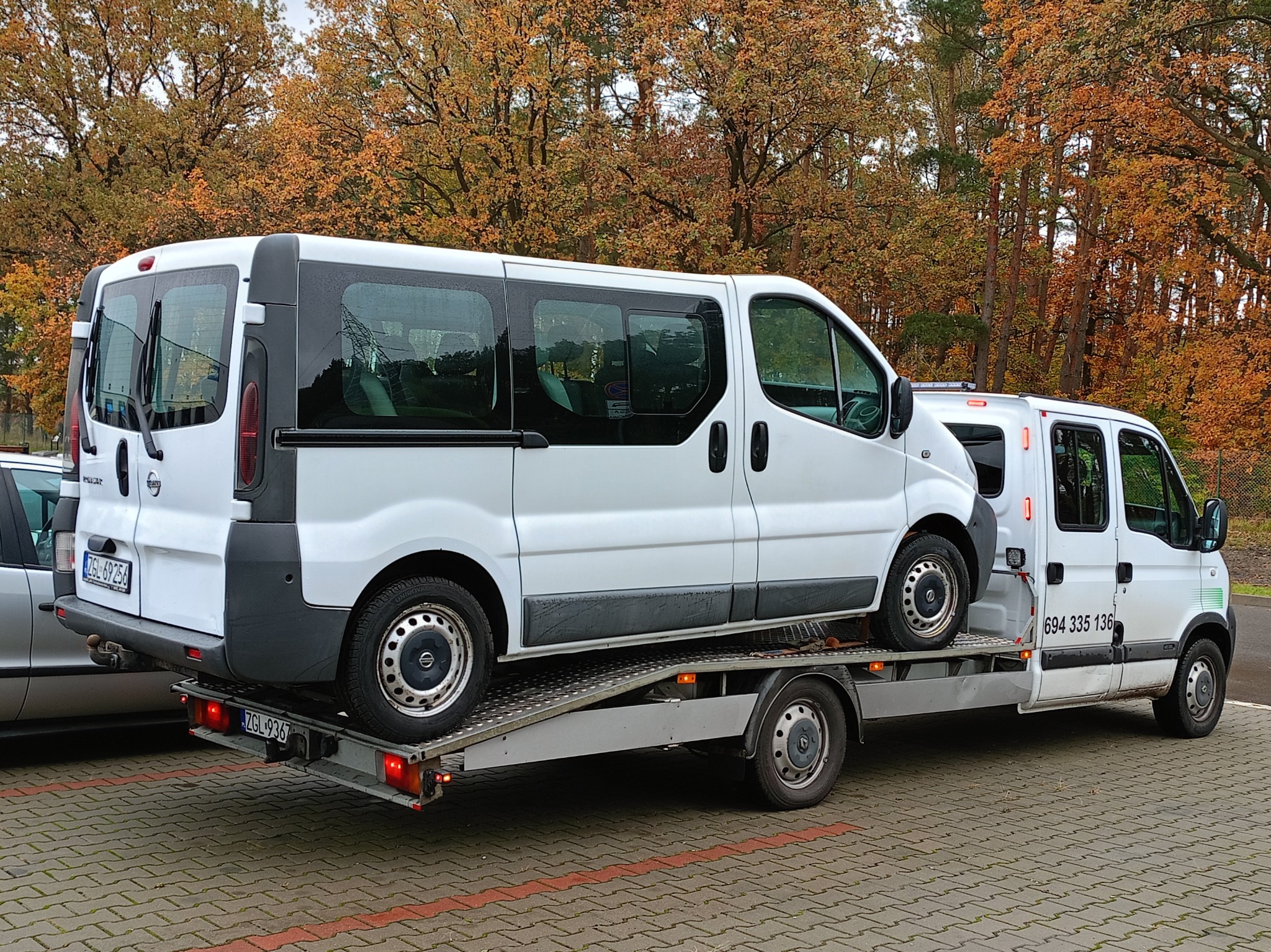 Biały bus na platformie holowniczej, widoczne numery rejestracyjne obu pojazdów, jesienne drzewa w tle.