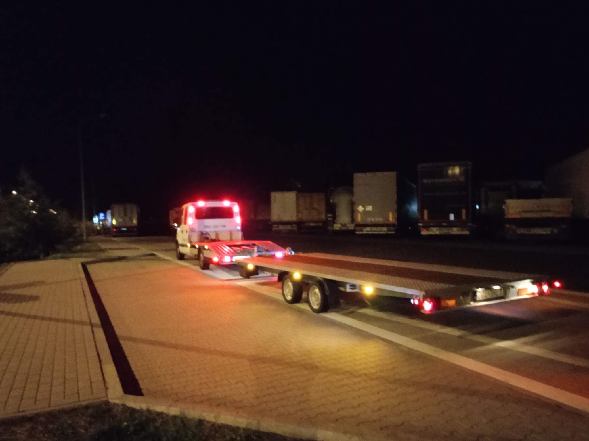 Ciężarówka z platformą transportową oświetlona światłami pozycyjnymi, stojąca na parkingu w nocy; w tle zaparkowane inne pojazdy ciężarowe.