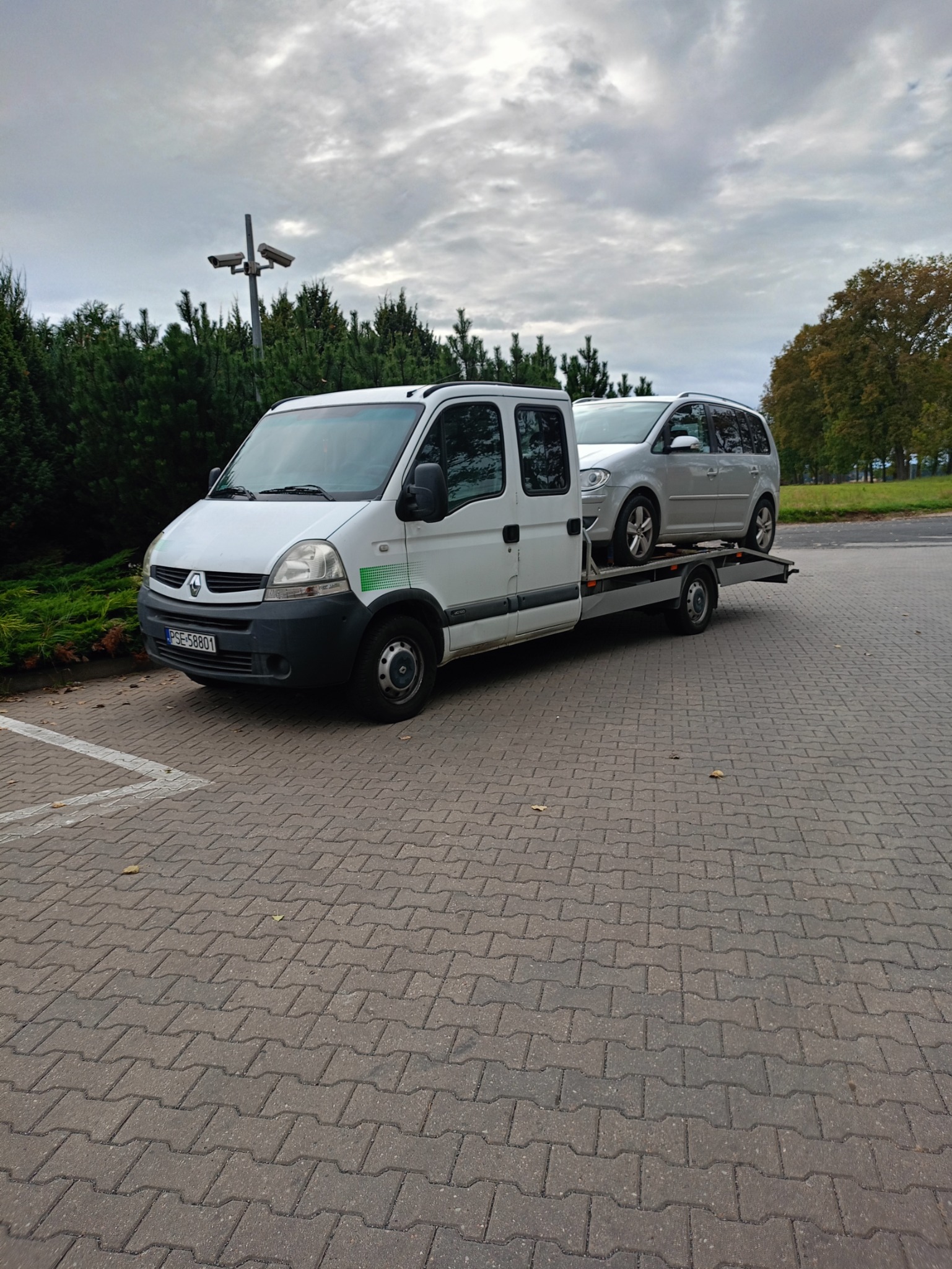 Biała platforma transportowa Renault z zielonymi elementami graficznymi przewozi srebrny samochód osobowy na parkingu z kostki brukowej, w tle widoczne drzewa i słup z kamerami monitoringu.