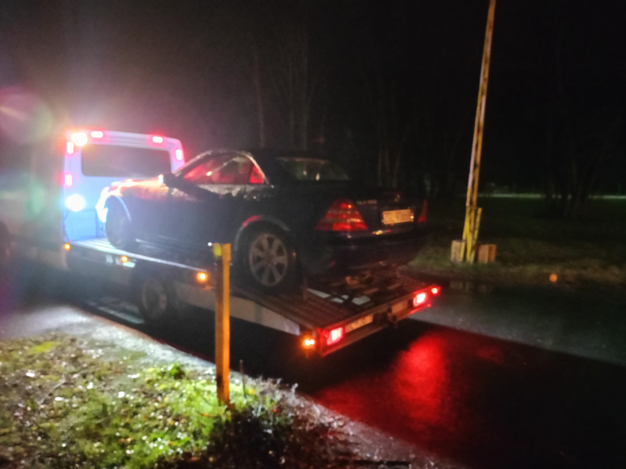 Czarny kabriolet Mercedes-Benz na platformie pojazdu pomocy drogowej w nocy, oświetlony światłami pojazdu, widoczne mokre podłoże i słupki barierki.