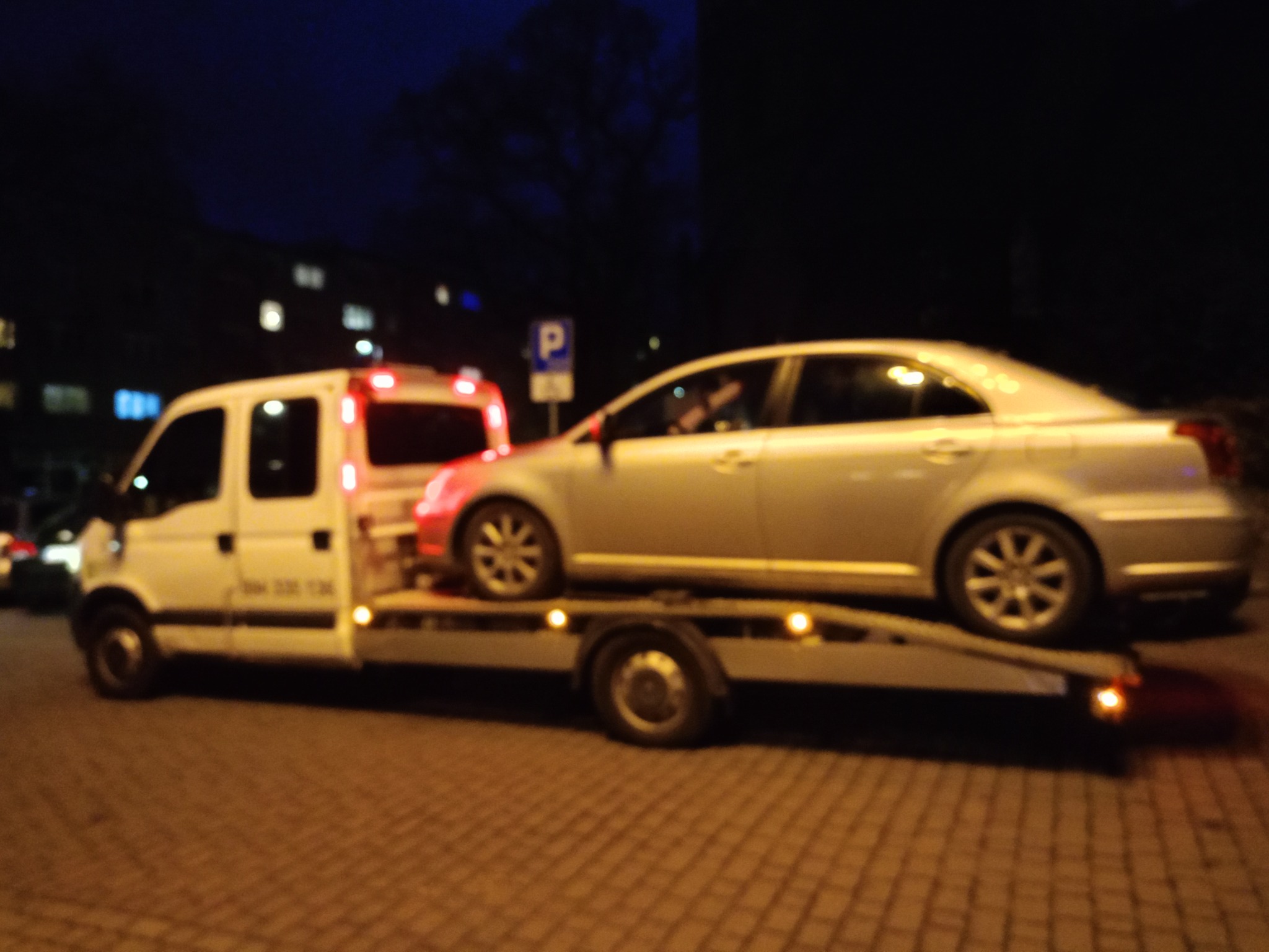 Srebrny samochód osobowy przewożony na platformie białego pojazdu pomocy drogowej, widoczne światła ostrzegawcze w nocy, na tle budynki i znak parkingu.