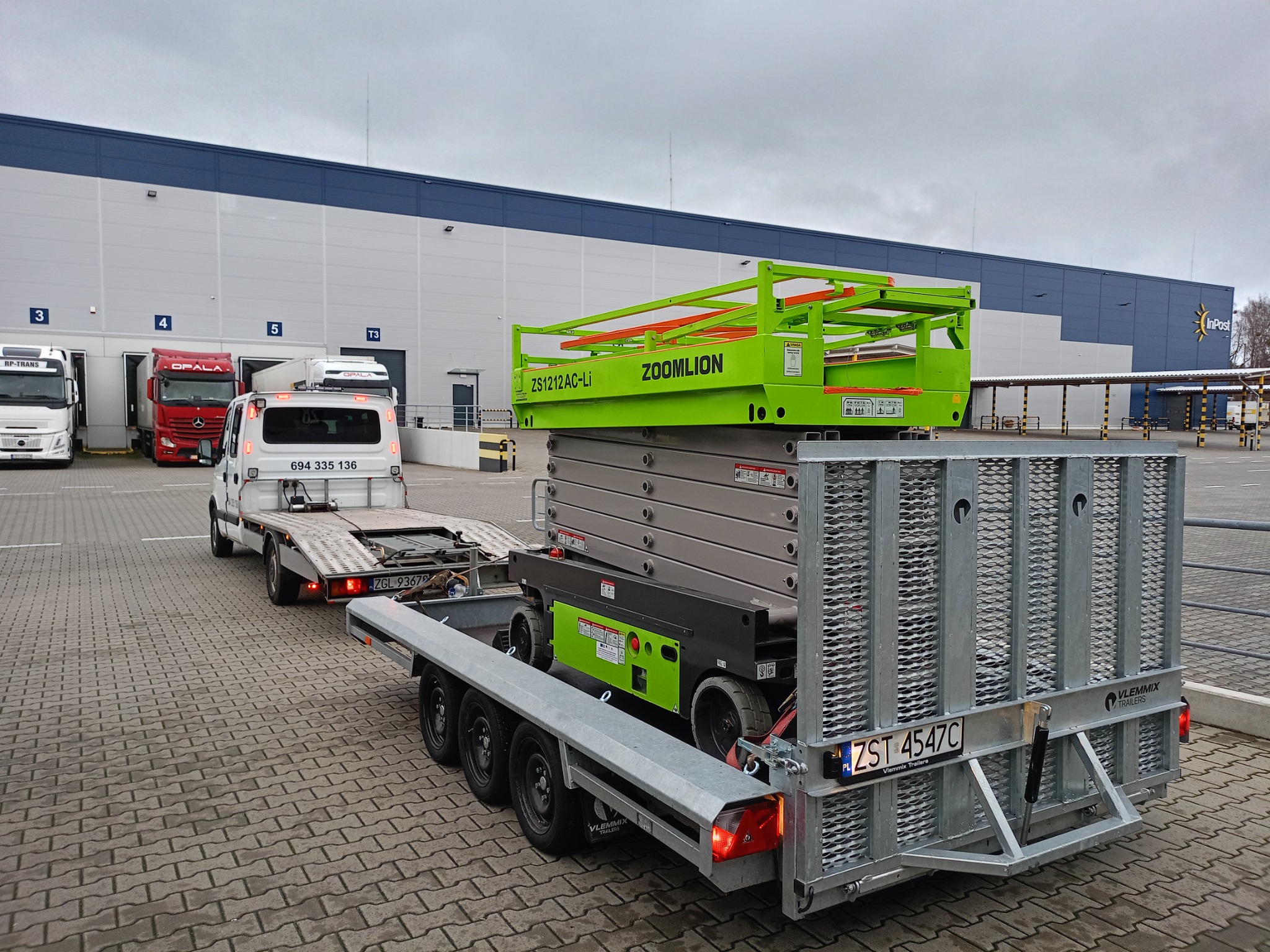 Transport platformy podnośnikowej Zoomlion ZS1212AC-Li na specjalistycznej przyczepie Vlemmix, z widocznym numerem rejestracyjnym ZST 4547C, na tle magazynu InPost.