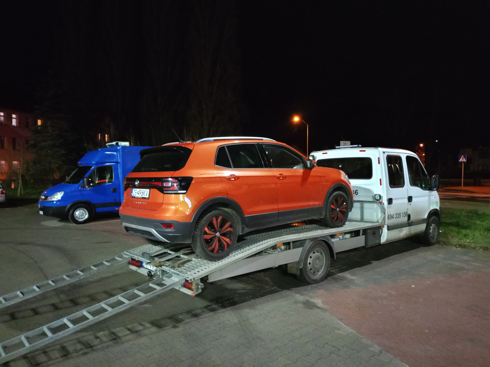 Pomarańczowy samochód osobowy z czarno-czerwonymi felgami transportowany na platformie pomocy drogowej podczas nocnej akcji, w tle zaparkowany niebieski bus.
