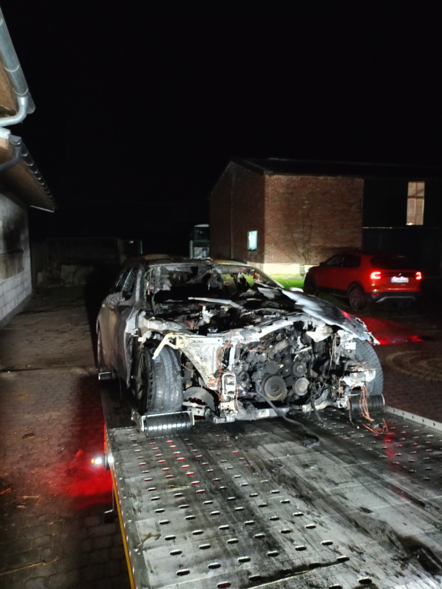 Spalony wrak samochodu osobowego bez karoserii, przewożony na platformie pomocy drogowej w nocnych warunkach, z czerwonym autem w tle.