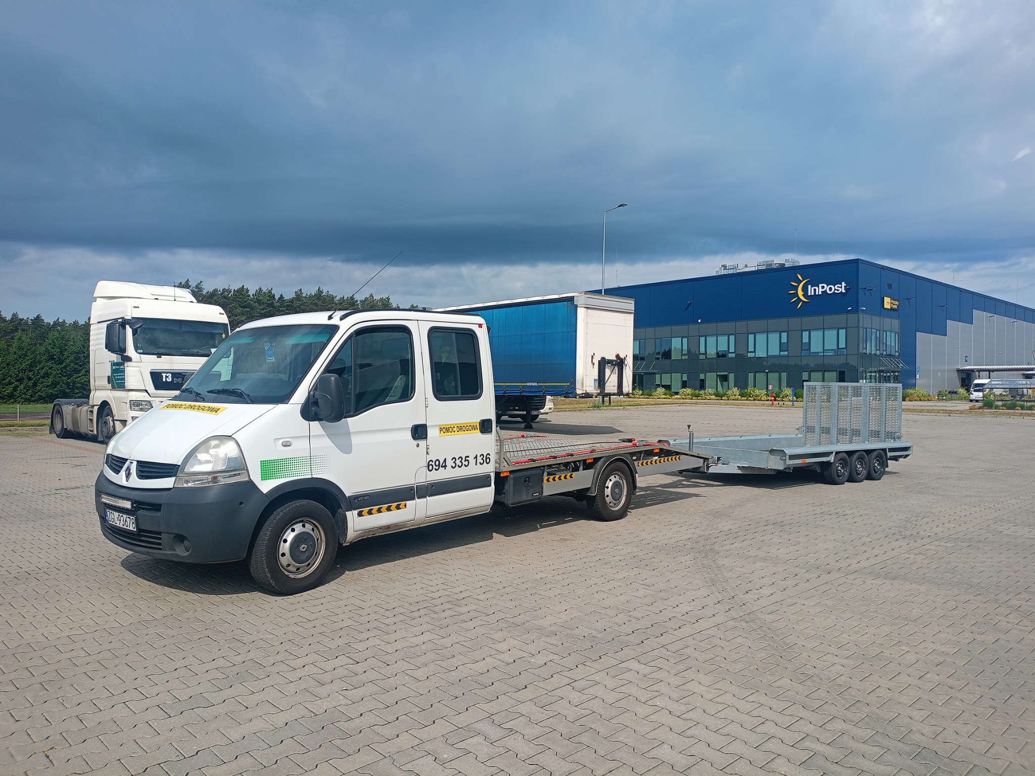 Biały bus pomocy drogowej z przyczepą na parkingu, w tle budynek InPost i ciężarówka. Napis 'Pomoc Drogowa' i numer telefonu na pojeździe.