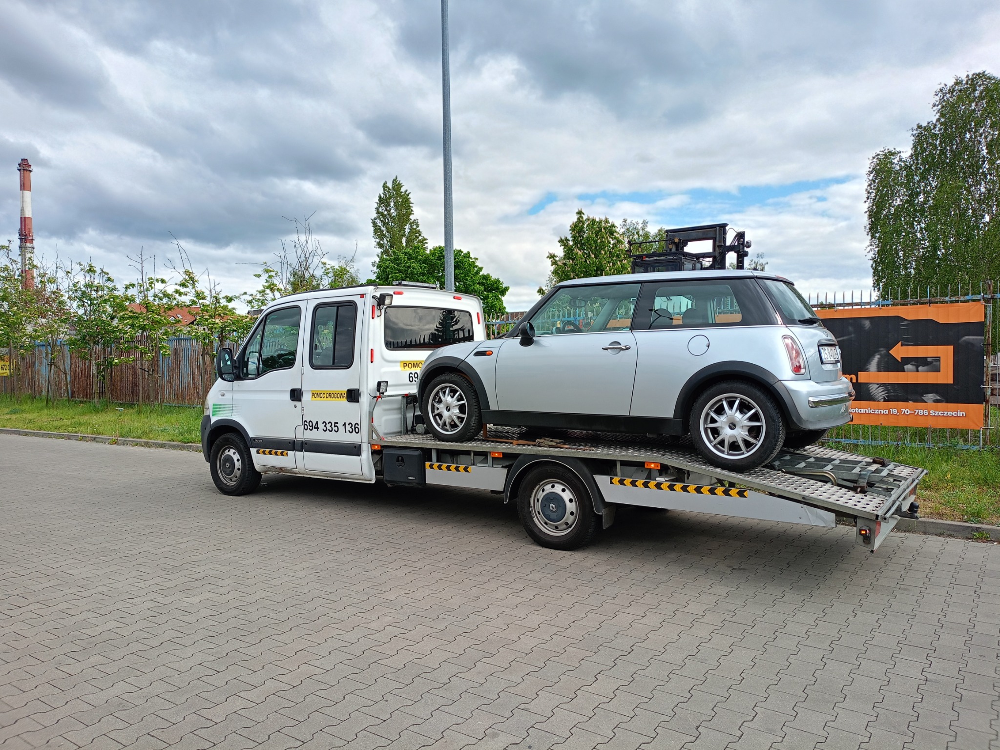 Laweta z Mini Cooperem na platformie, gotowa do transportu w Goleniowie. Widoczny numer telefonu do pomocy drogowej i adres firmy.