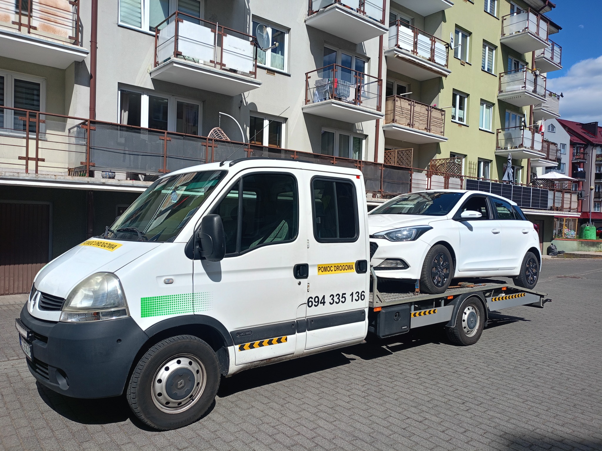 Biała laweta Renault z numerem telefonu 694 335 136 przewozi biały samochód osobowy na tle budynku mieszkalnego.