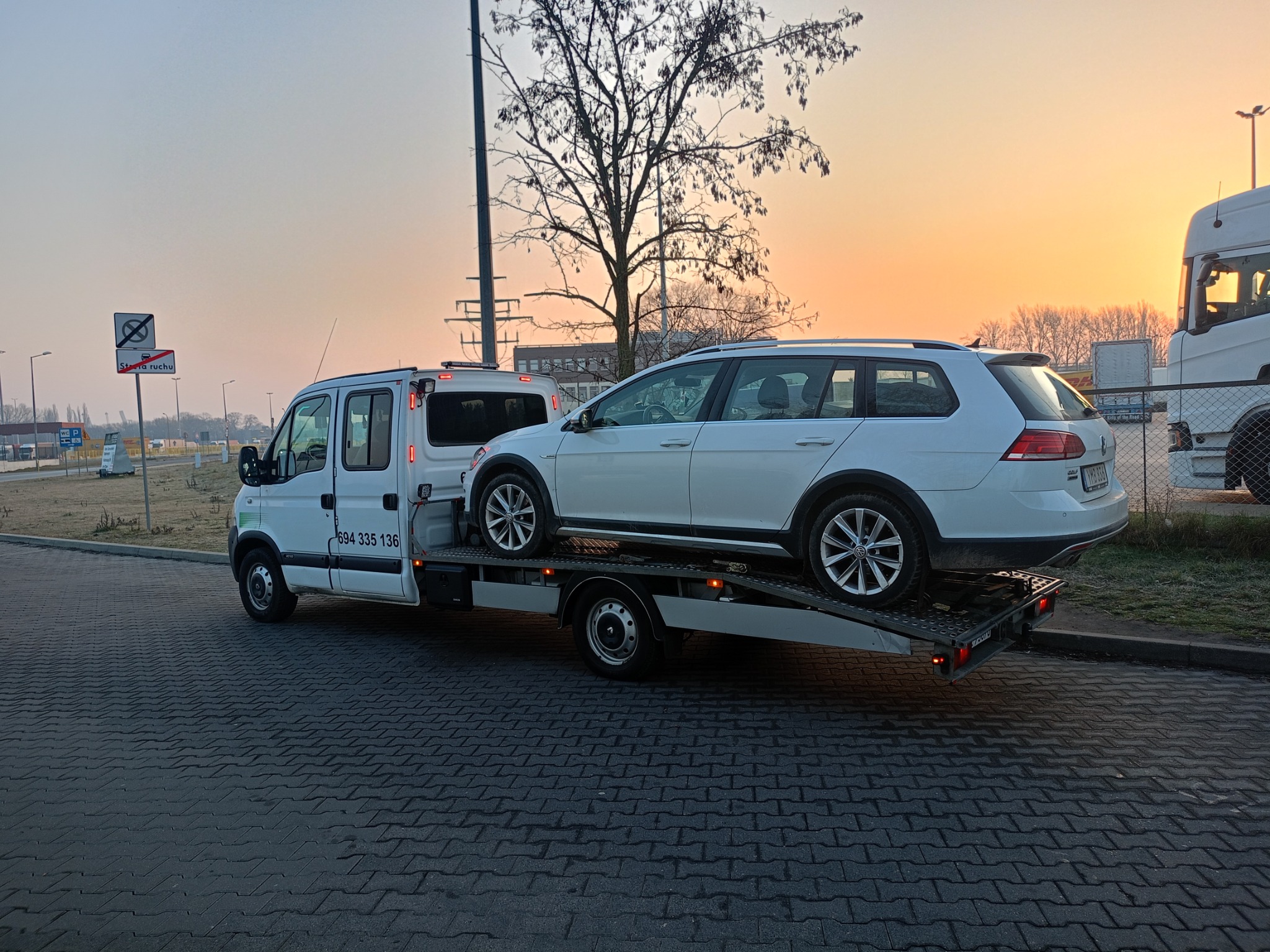 Laweta z białym VW Golf Alltrack na platformie, zaparkowana na brukowanej drodze, widok z boku, w tle budynki i drzewa o zachodzie słońca.