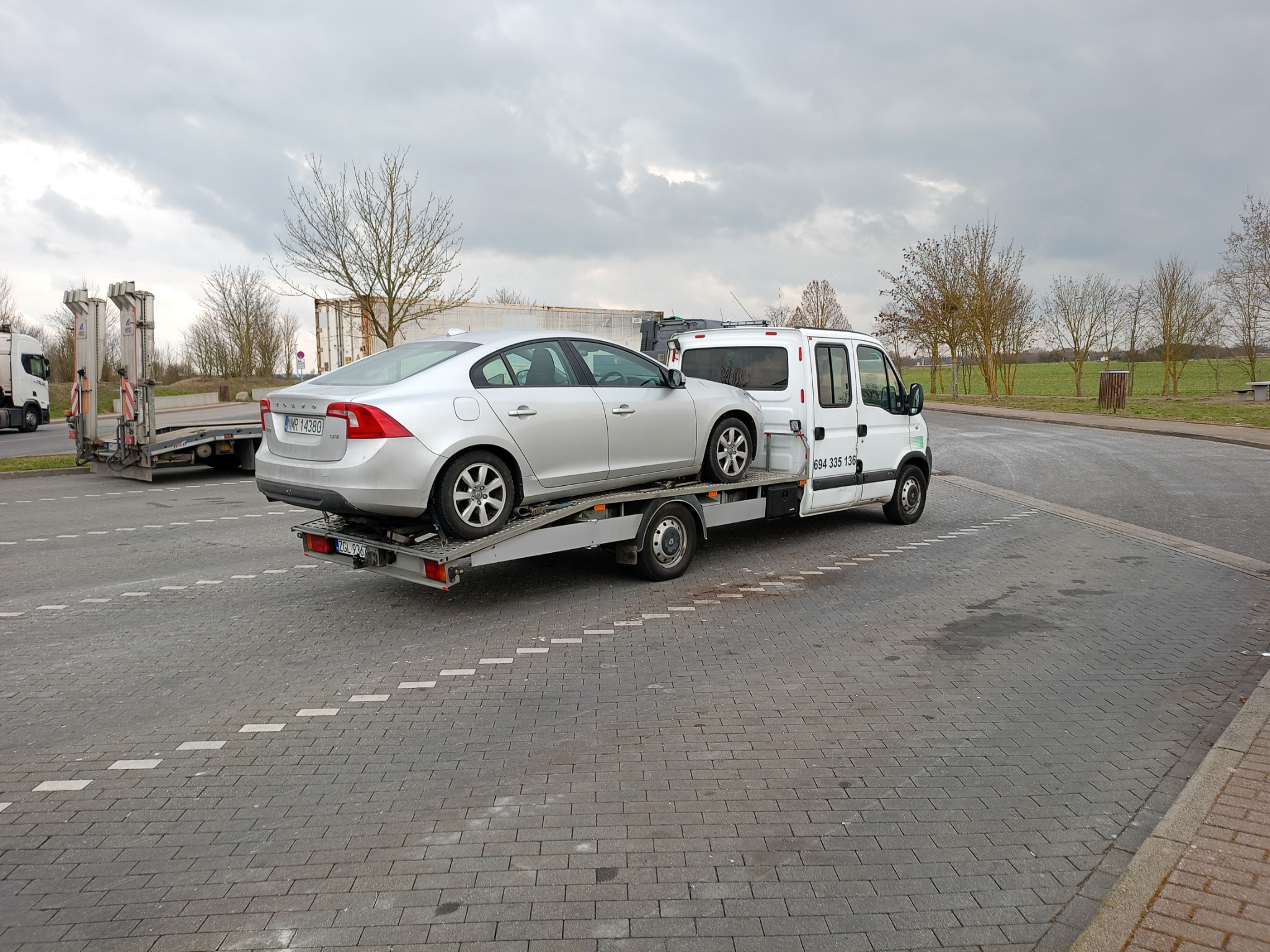 Srebrny samochód osobowy Volvo na platformie lawety holowany przez biały pojazd dostawczy na parkingu z kostki brukowej w pochmurny dzień.