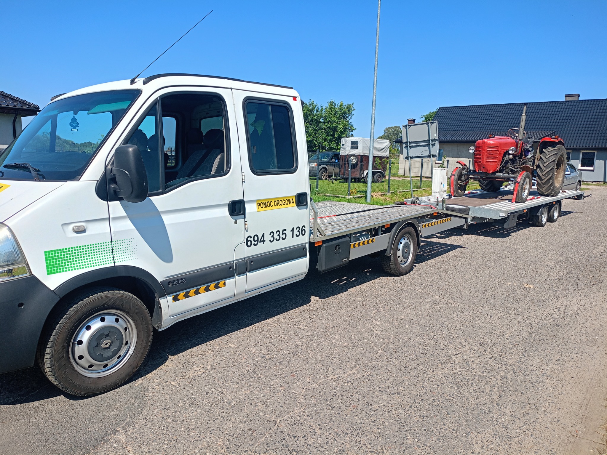 Laweta z czerwonym traktorem na platformie. Biały bus z napisem 'Pomoc Drogowa' i numerem telefonu. Transport maszyny rolniczej w słoneczny dzień.